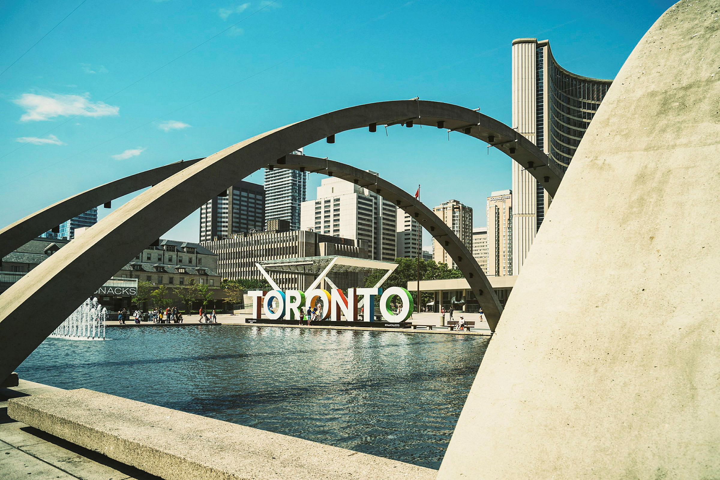 Näkymä Nathan Phillips Squaresta Torontossa, jossa suihkulähde, kaaret ja kaupungin siluetti taustalla. (Tekoälyn luoma teksti)