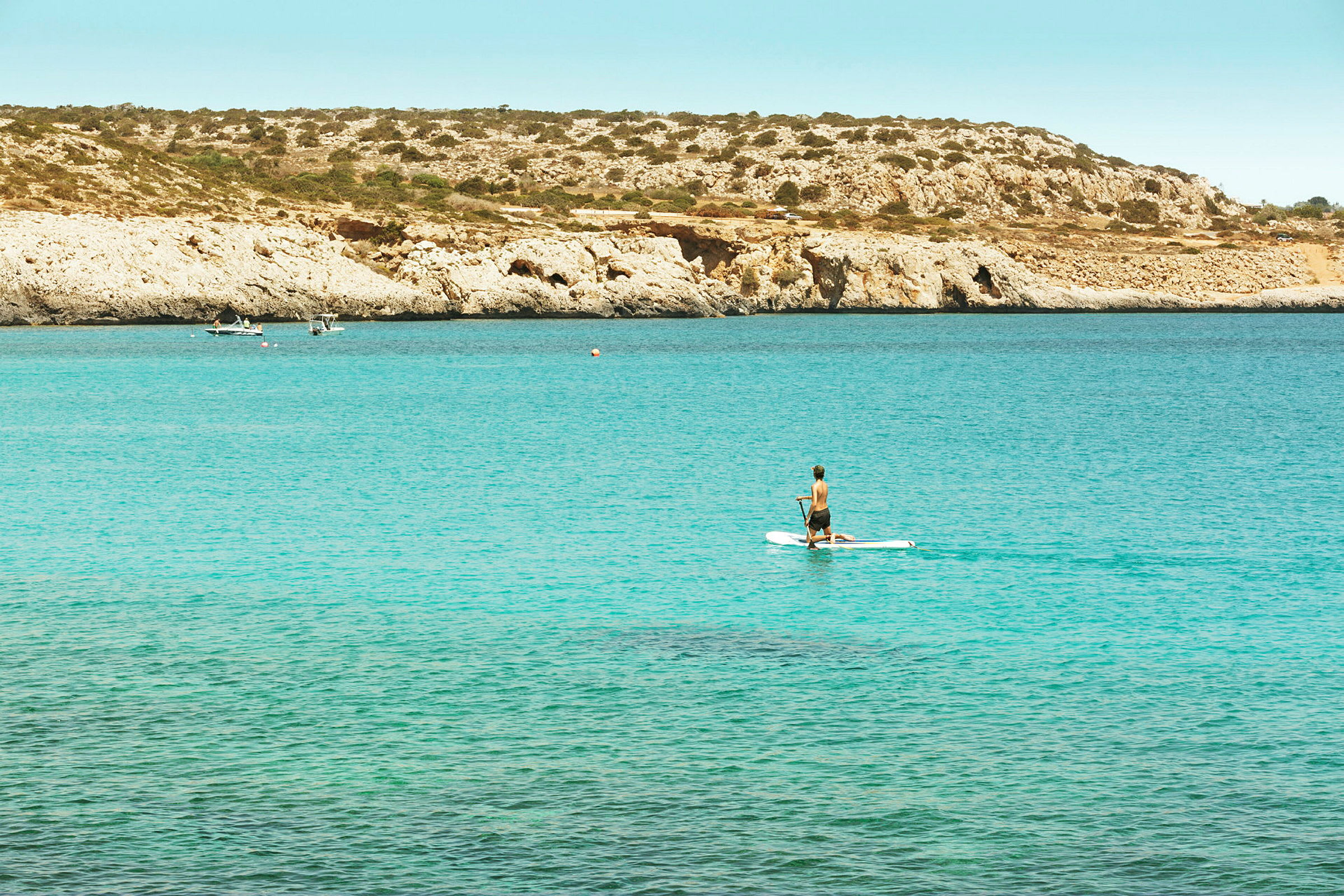 Henkilö meloo paddleboardilla taustalla kallioita ja merta. (Tekoälyn luoma teksti)