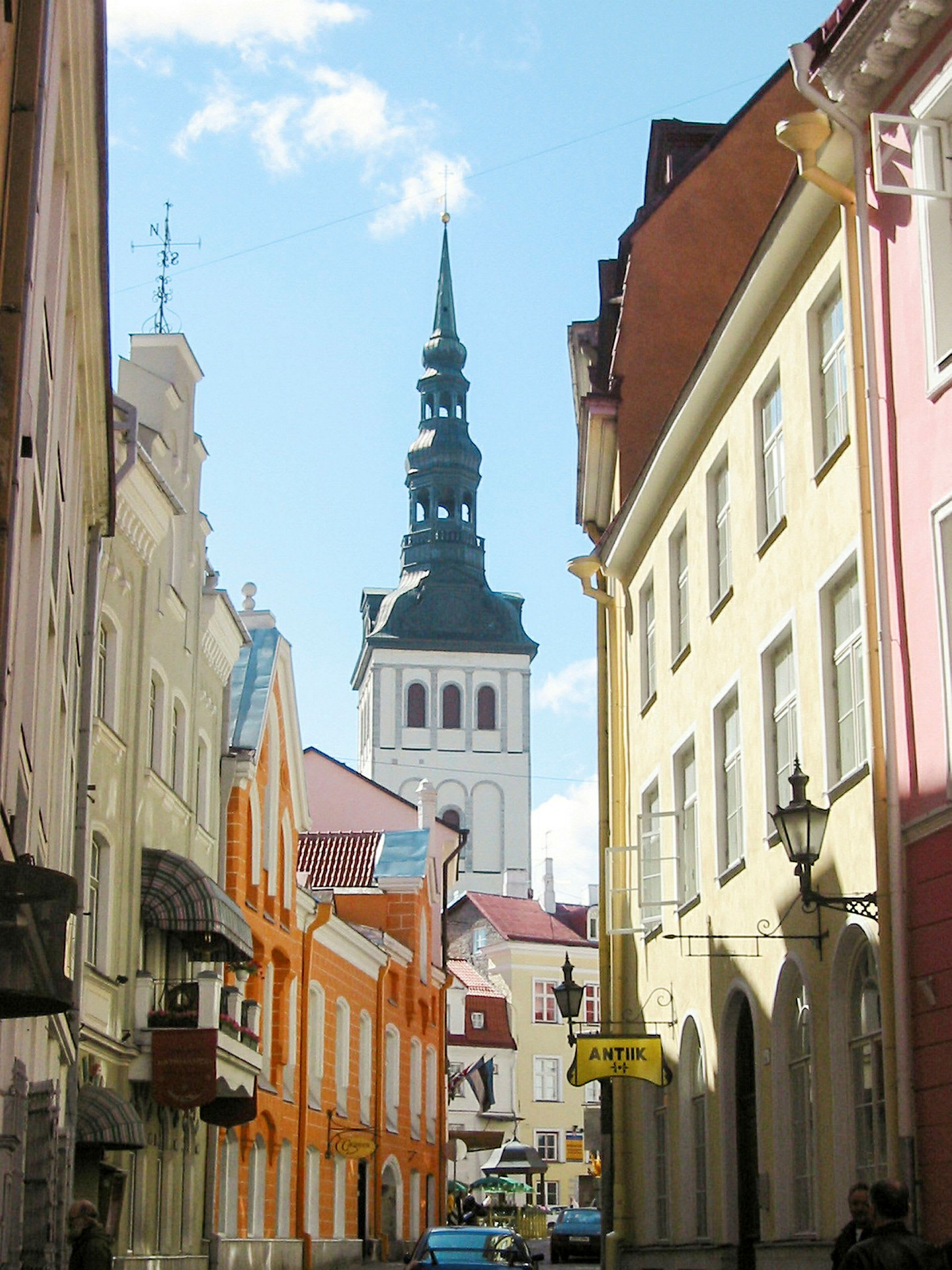 Kapea katu, jota reunustavat värikkäät rakennukset, ja korkea kirkontorni taustalla. (Tekoälyn luoma teksti)