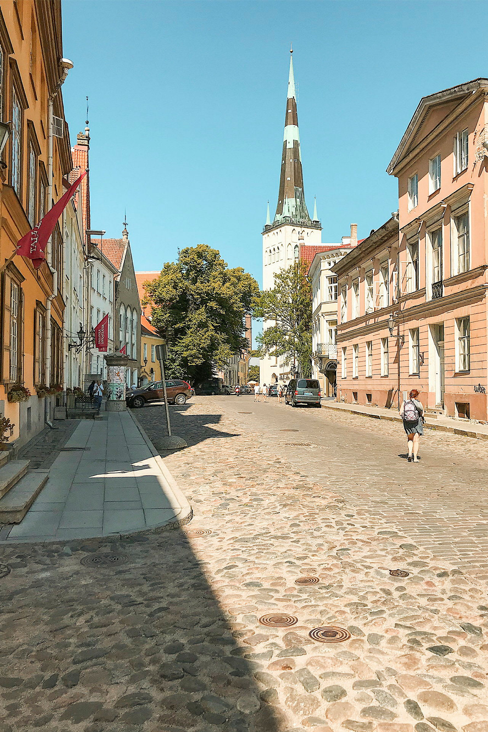 Kivipäällysteinen katu, jota reunustavat värikkäät vanhat rakennukset, ja taustalla näkyy kirkontorni kirkkaan sinisen taivaan alla. (Tekoälyn luoma teksti)