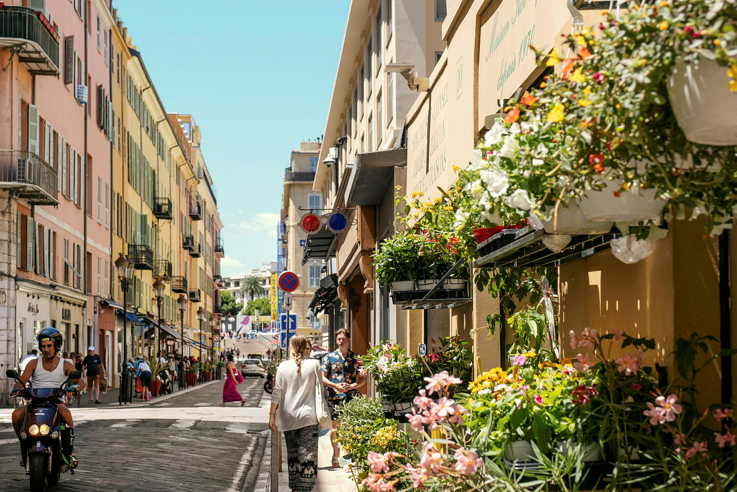 Kaupunkikatu, jonka rakennukset ovat raidallisen värikkäitä. Julkisivulla roikkuu kukkia, ja ihmiset kävelevät ja ajavat mopolla kadulla. (Tekoälyn luoma teksti)