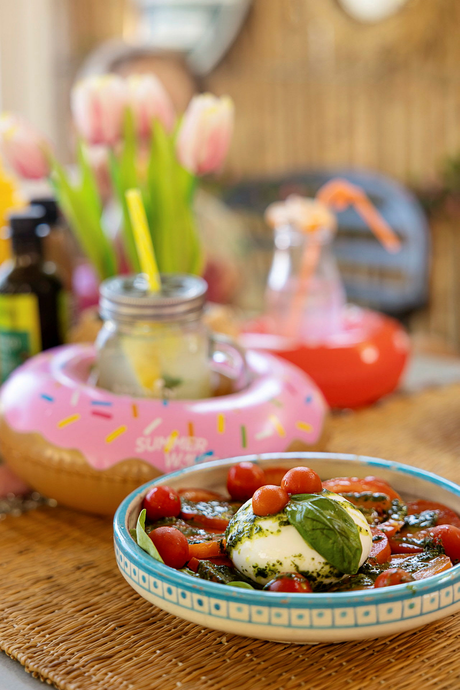 Ruokalaji, jossa on buffalo-mozzarellaa, tomaatteja ja basilikaa sekä koristeltuja juomia. Taustalla näkyy tulppaaneja ja muita koriste-esineitä. (Tekoälyn luoma teksti)
