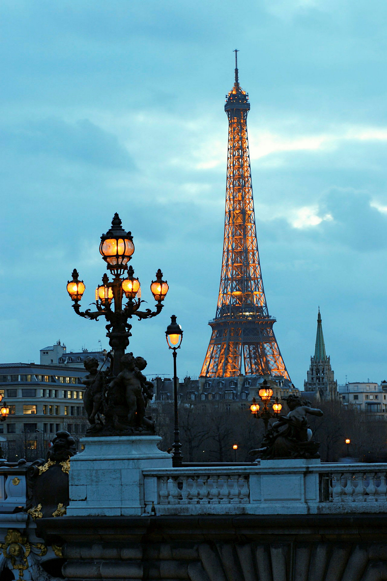 Eiffel-torni valaistuna hämärässä, kaupungin siluetti ja koristeelliset katuvalot etualalla. (Tekoälyn luoma teksti)