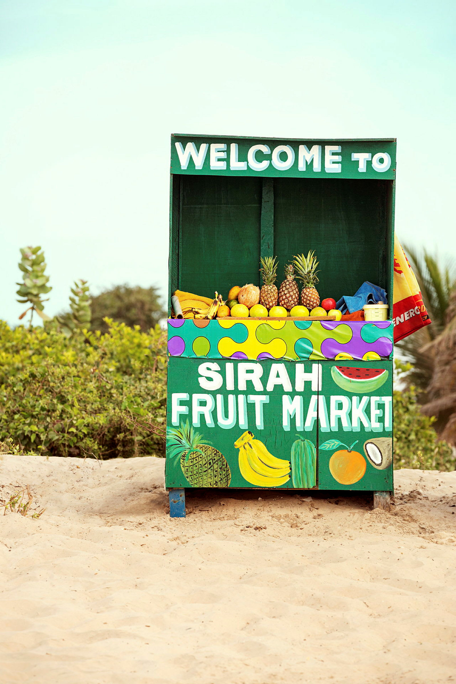 Hedelmäkoju, jossa lukee "Sirah Fruit Market", tarjoaa erilaisia hedelmiä, kuten ananaksia, banaaneja ja sitrushedelmiä. Koju sijaitsee hiekkamaalla ja taustalla on kasvillisuutta. (Tekoälyn luoma teksti)