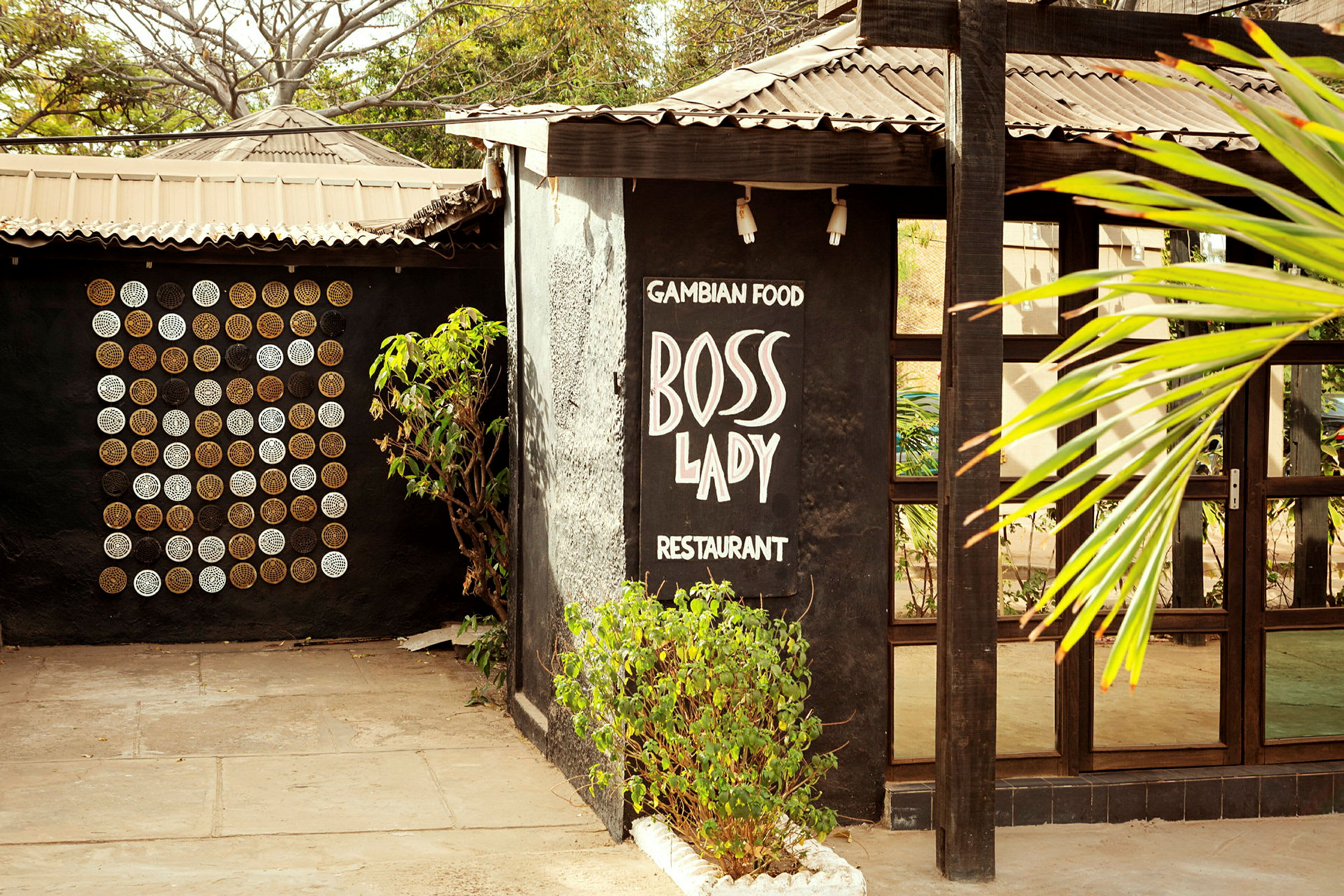 Ravintola, jossa on teksti 'Gambian Food Boss Lady Restaurant' mustalla seinällä. Ympärillä on kasvillisuutta ja vasemmalla näkyy koristeellinen seinä ympyröineen. (Tekoälyn luoma teksti)