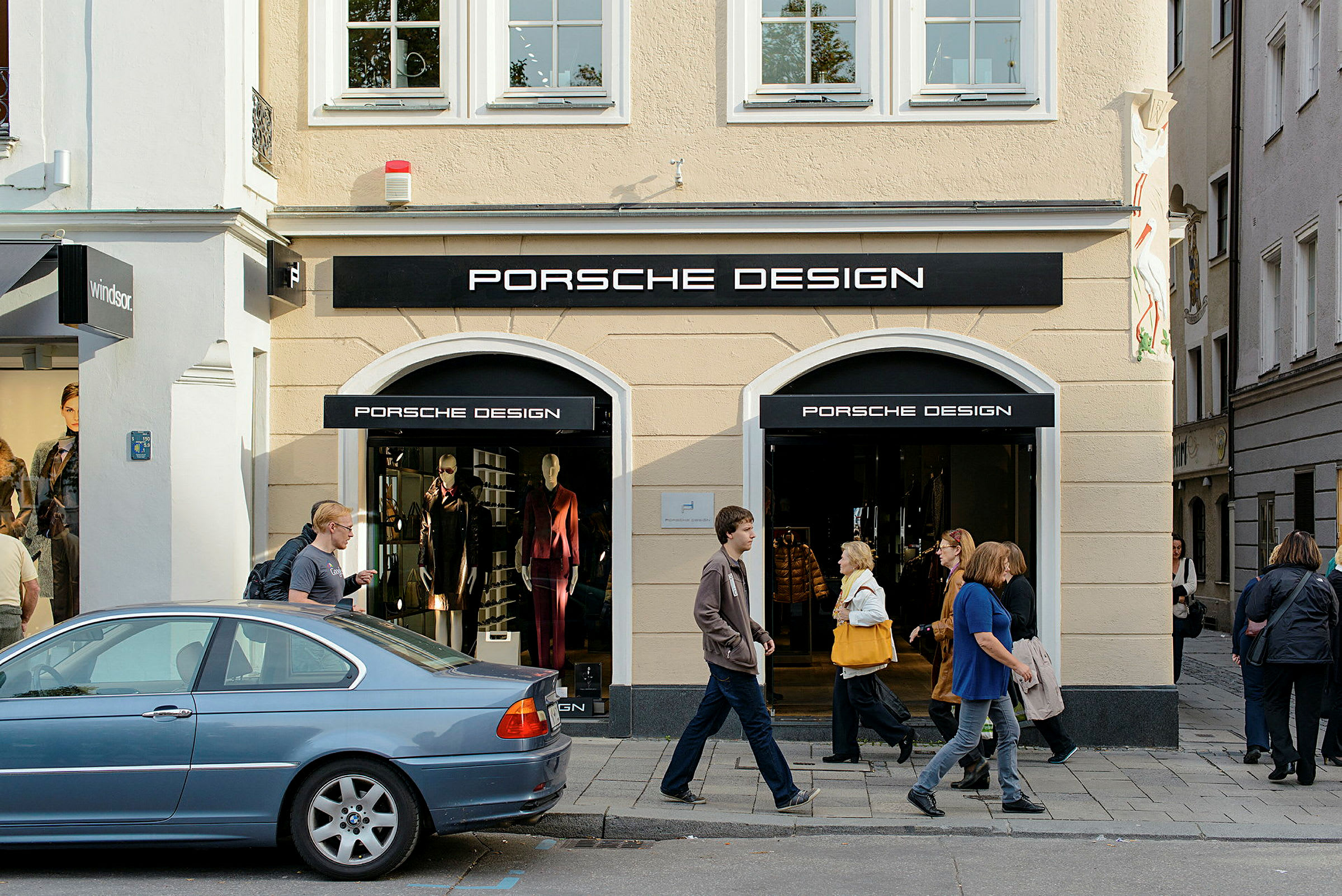 Sisäänkäynti kauppaan, jossa Porsche Design -logo, näyteikkunat ja ihmisiä kävelemässä ulkopuolella jalkakäytävällä. (Tekoälyn luoma teksti)