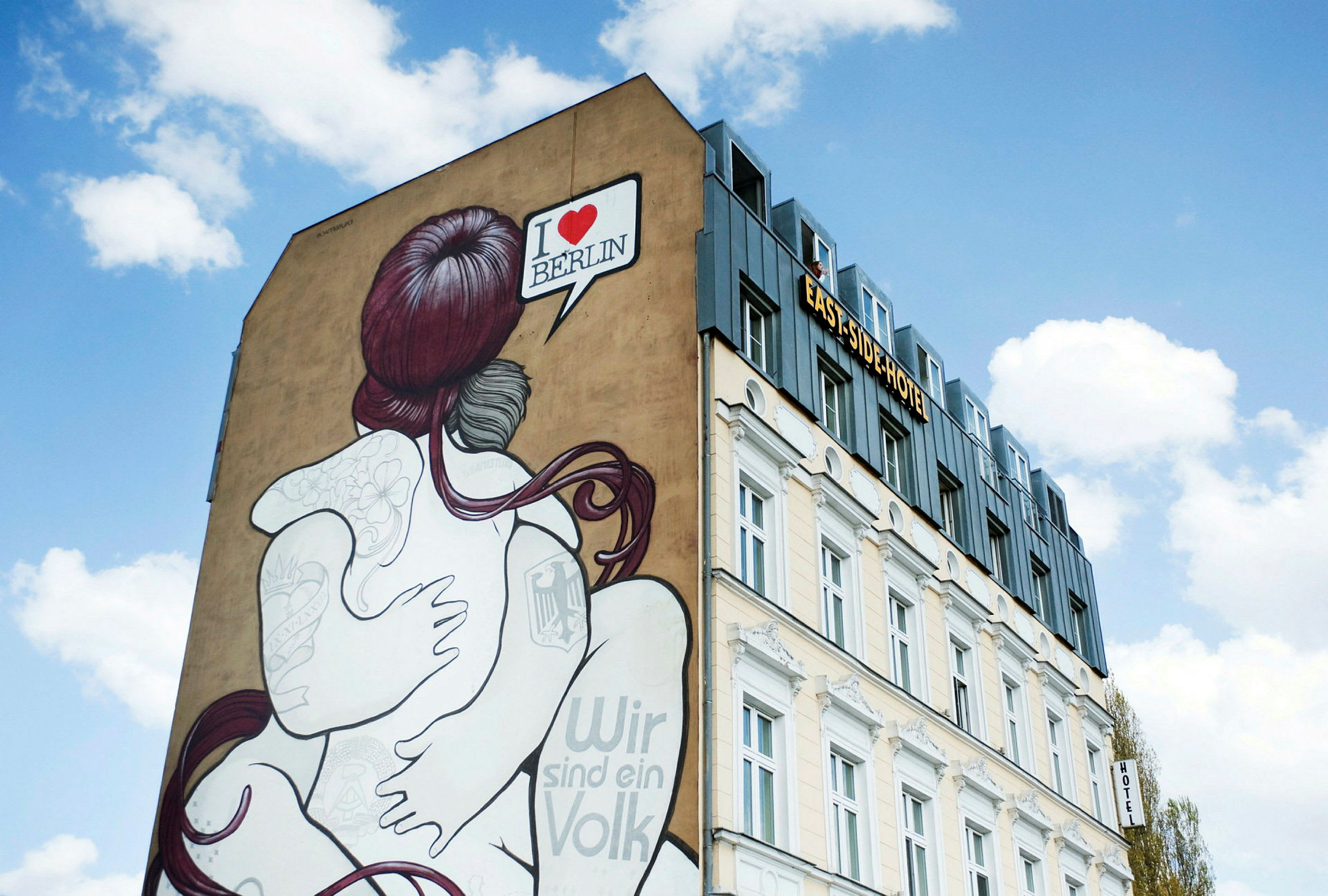 Rakennus, jonka seinämaalauksessa näkyy toisiaan syleilevä pari. Kuvassa on puhekupla, jossa lukee "I ♥ BERLIN". Taivas on kirkas valkoisine pilvineen. (Tekoälyn luoma teksti)