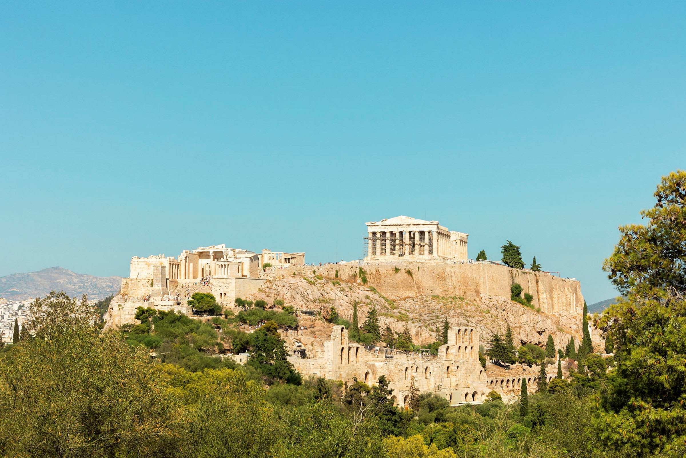 Valokuva Akropoliksesta, jonka huipulla on Parthenon, ympäröitynä vehreydellä ja taustalla kirkkaan sininen taivas. (Tekoälyn luoma teksti)