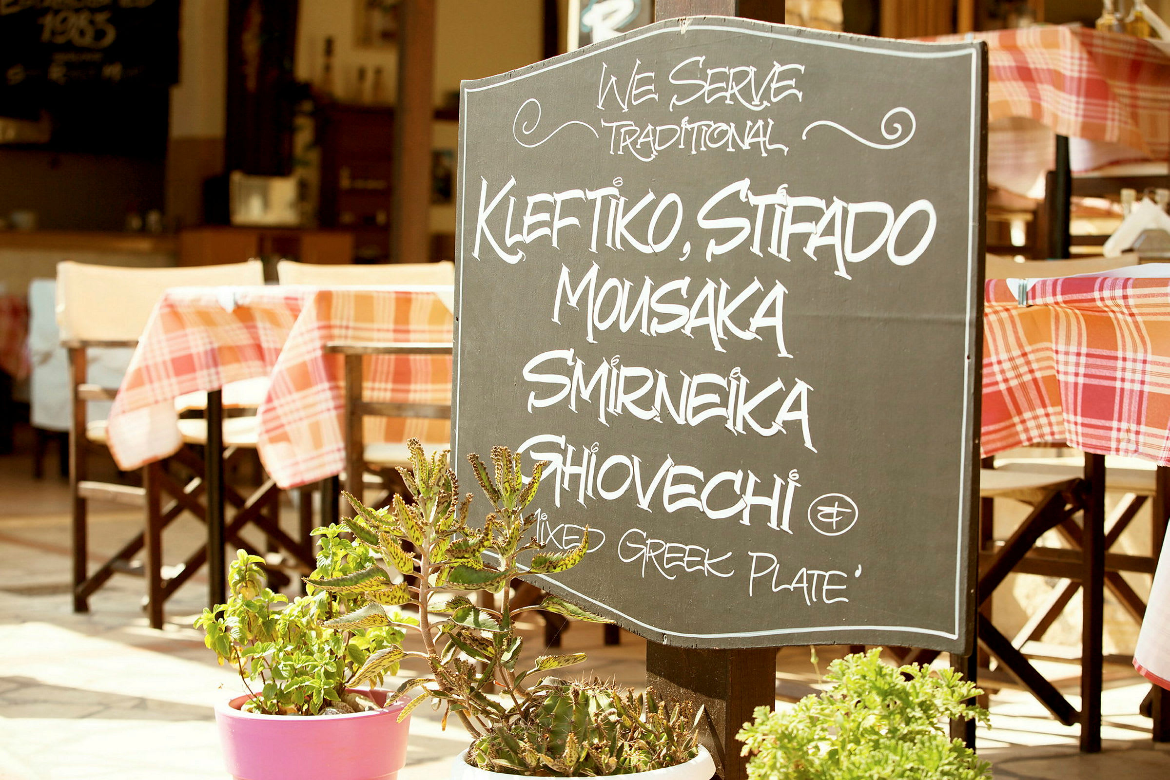 Ravintolan ulkopuolella oleva kyltti, jossa luetellaan perinteisiä kreikkalaisia ruokia. (Tekoälyn luoma teksti)