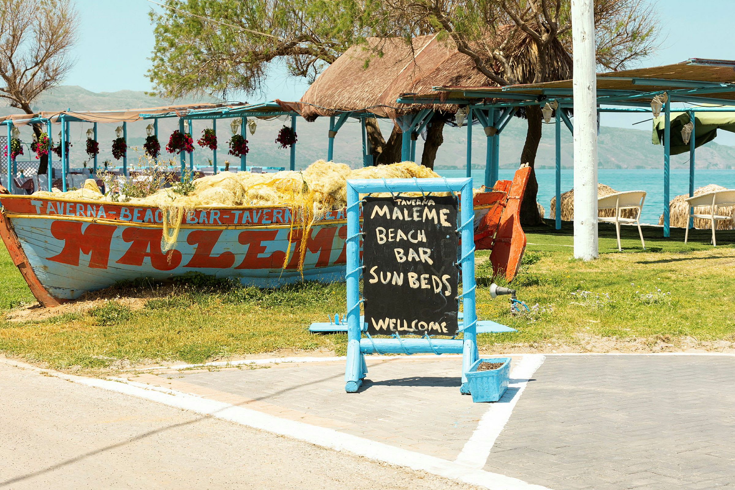 Kyltti, jossa lukee 'Maleme Beach Bar Sunbeds Welcome', vene koristeltu verkoilla ja ulkoterassi meren äärellä. (Tekoälyn luoma teksti)