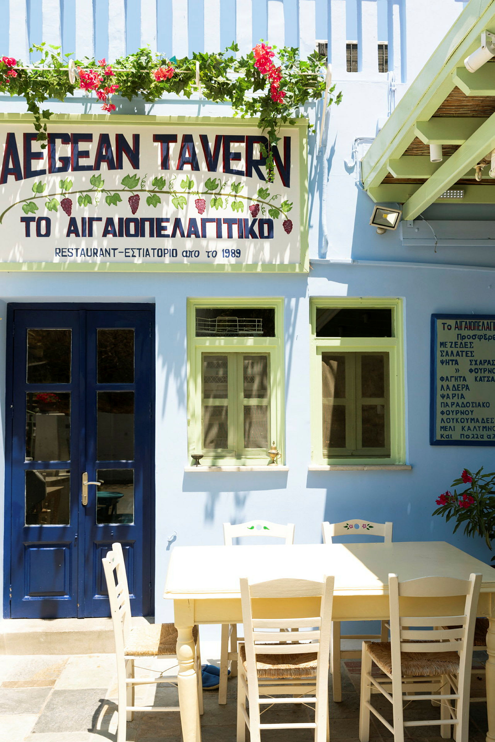 Rakennus, jossa on sininen ovi ja vihreät ikkunat, kyltti, jossa lukee 'AEGEAN TAVERN', ja pöytä tuoleineen edessä. (Tekoälyn luoma teksti)