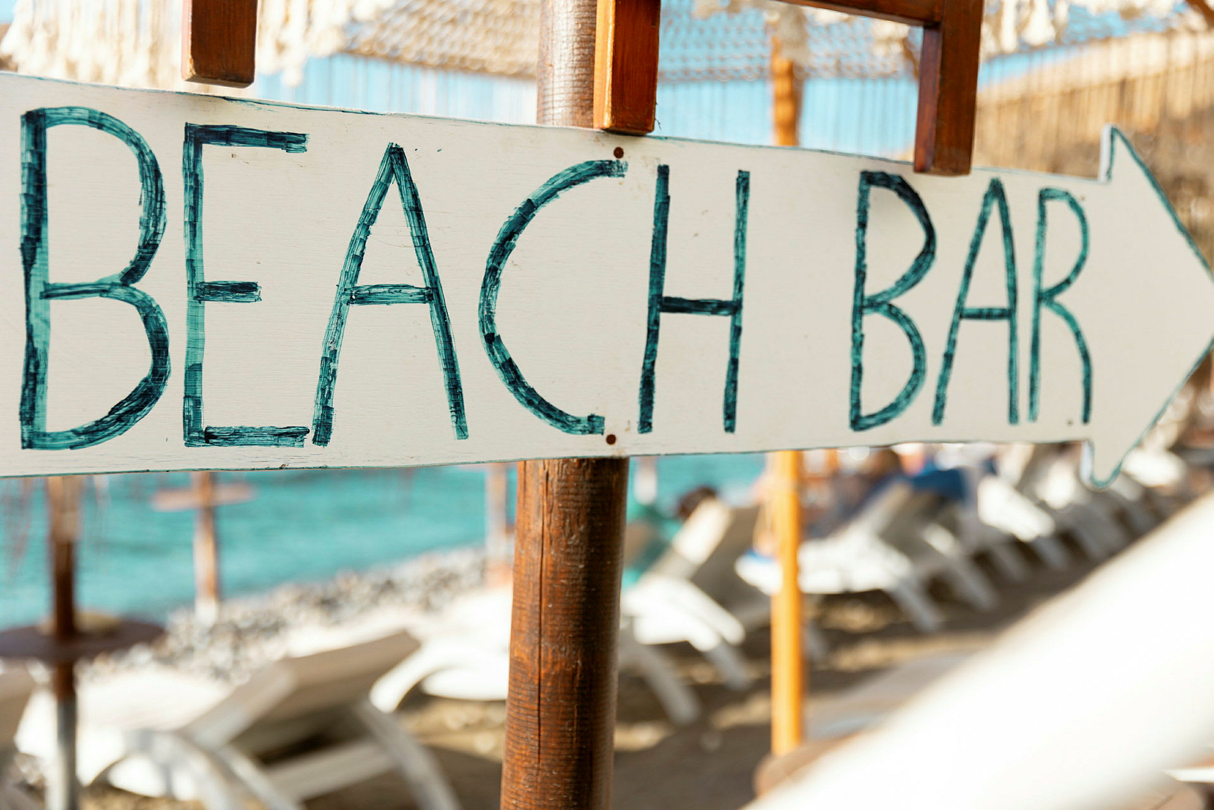Kyltti, jossa teksti 'BEACH BAR' ja nuoli, taustalla ranta aurinkotuoleineen ja meri. (Tekoälyn luoma teksti)