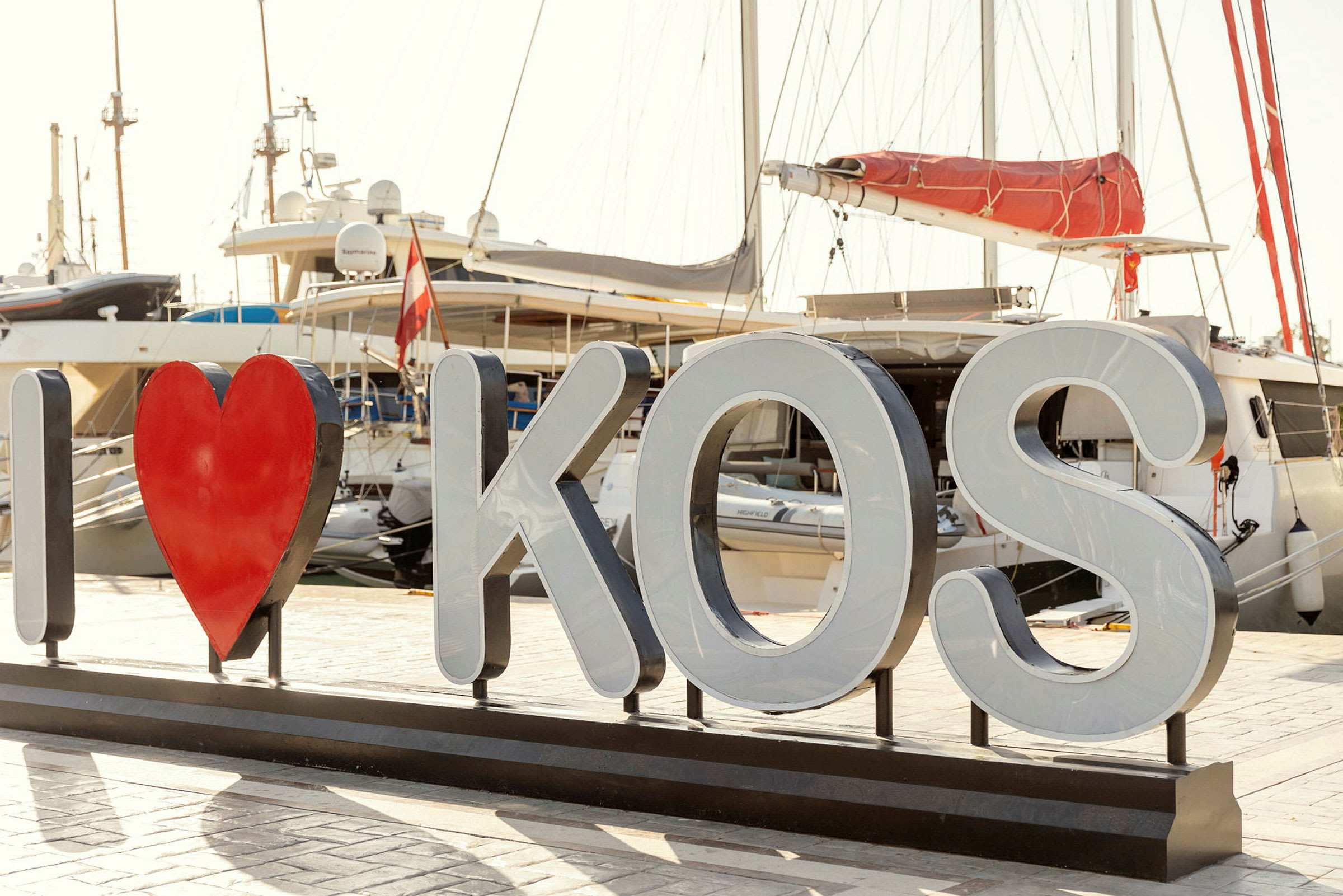 Kyltti, jossa teksti 'I ♥ KOS' veneiden edessä satamassa. (Tekoälyn luoma teksti)