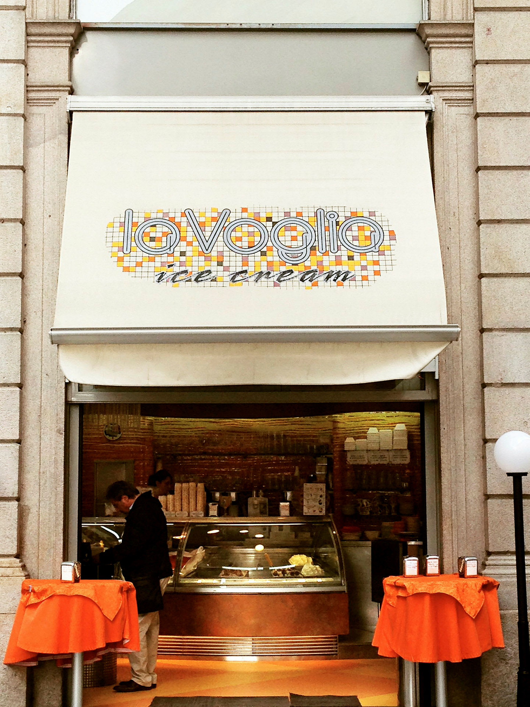 Jäätelökauppa, jossa on kyltti "La Voglia ice cream" ja kaksi pientä pöytää oransseilla liinoilla sisäänkäynnin edessä. (Tekoälyn luoma teksti)