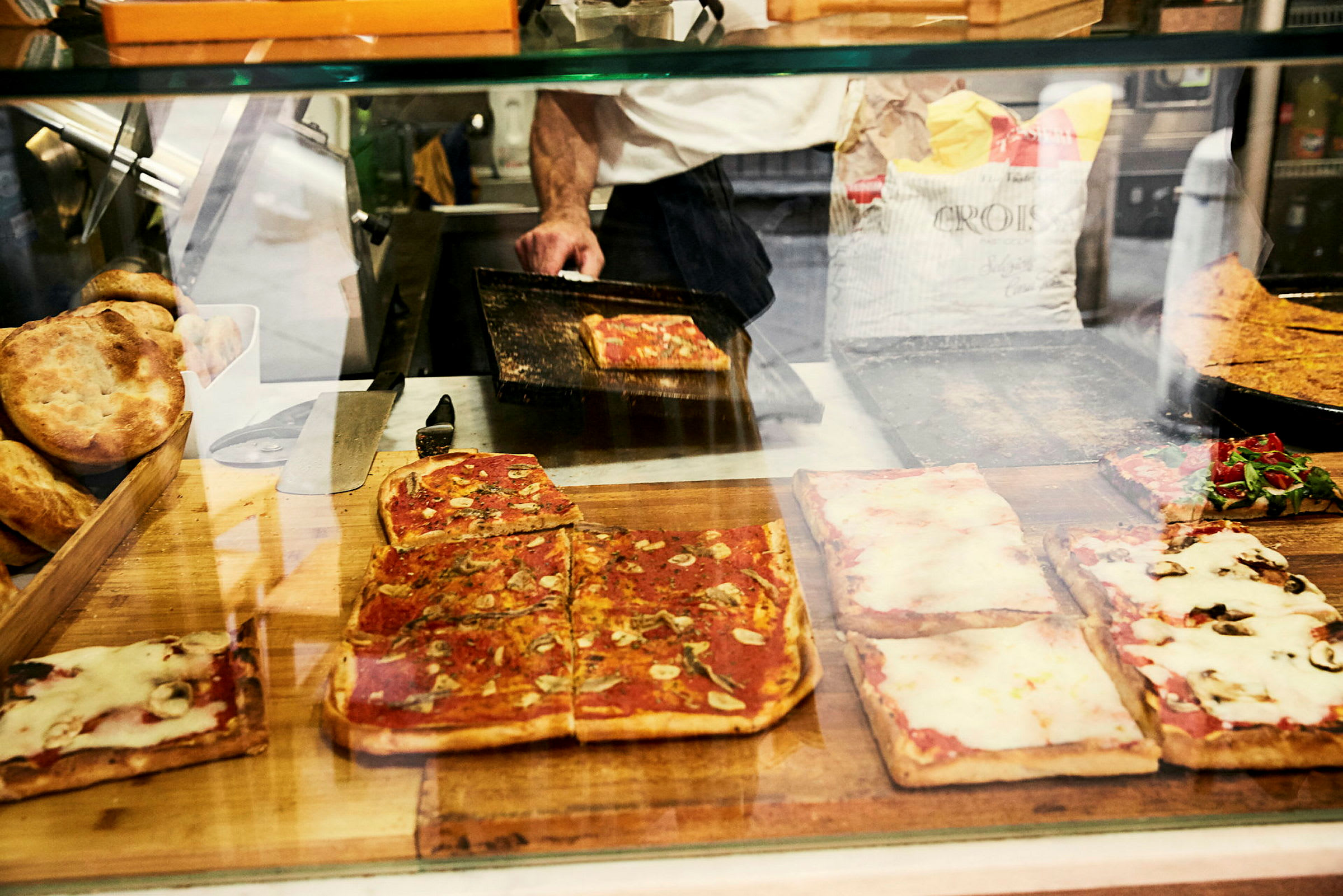 Pizzapaloja, joissa erilaisia täytteitä lasin takana, sekä leipää ja pizzauuni taustalla. (Tekoälyn luoma teksti)