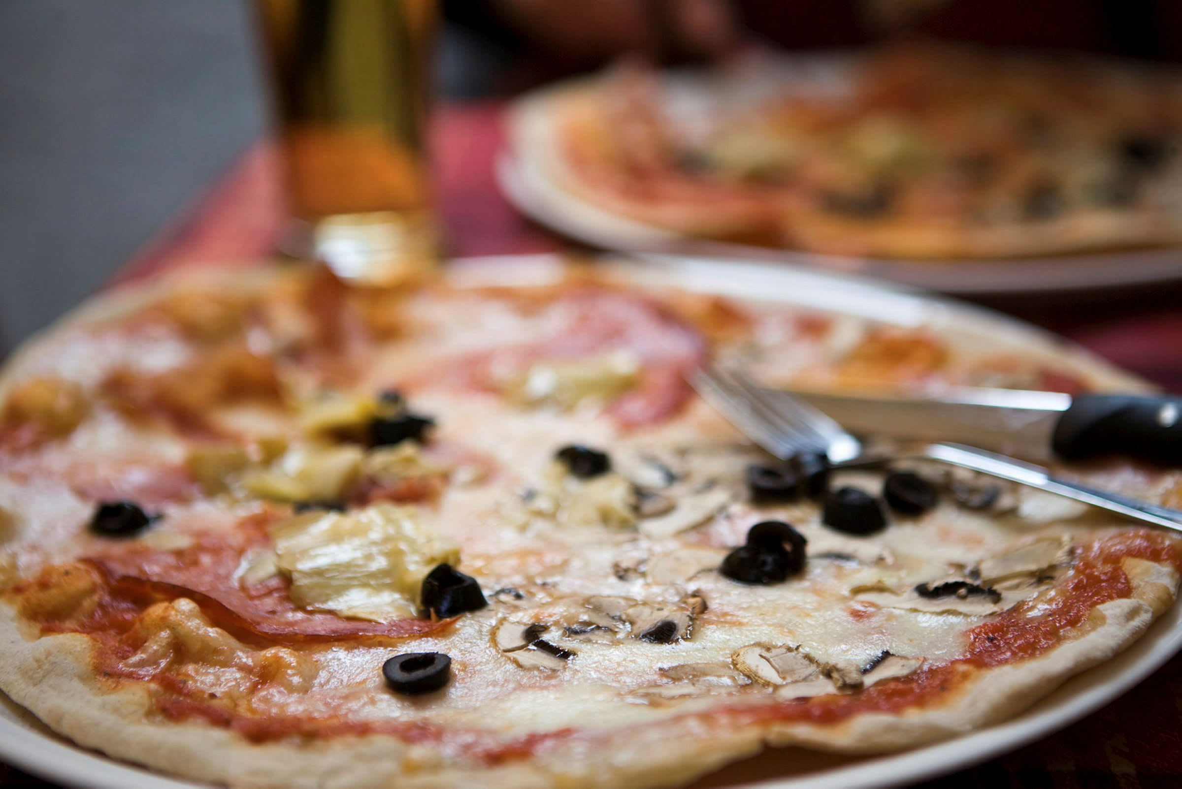Lautanen, jossa pizzaa, päällä juustoa, mustia oliiveja ja herkkusieniä. Aterimet ovat lautasen reunalla. (Tekoälyn luoma teksti)