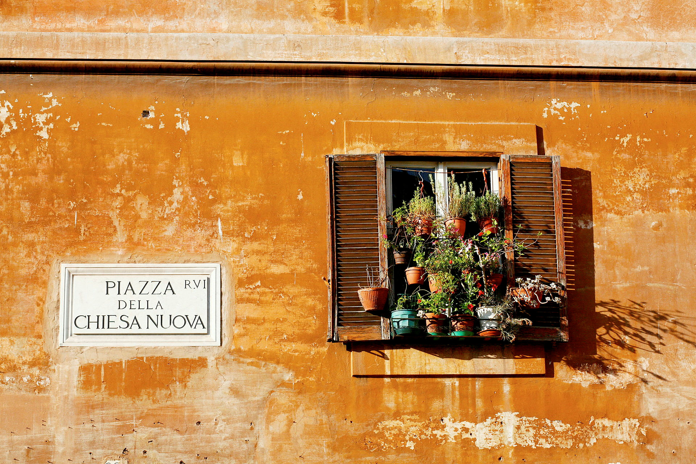 Ikkuna, jossa kukkaruukkuja keltaiseksi maalatulla seinällä, kyltin vieressä, jossa lukee 'Piazza della Chiesa Nuova'. (Tekoälyn luoma teksti)