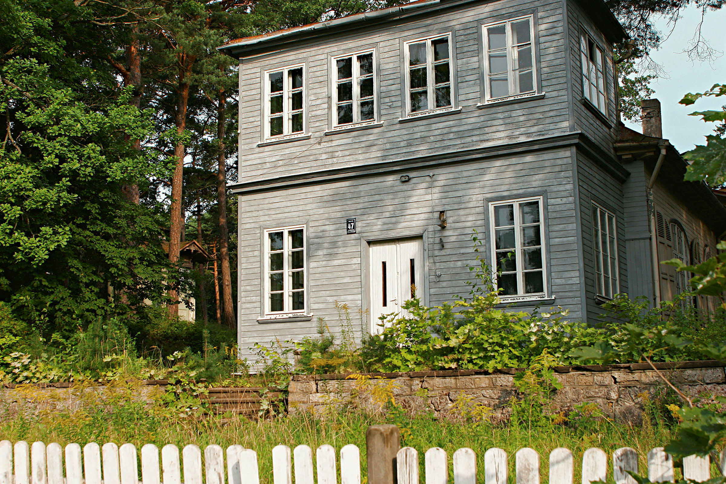 Kaksikerroksinen harmaa puutalo, jota ympäröi vehreys ja jonka etualalla on valkoinen aita. (Tekoälyn luoma teksti)
