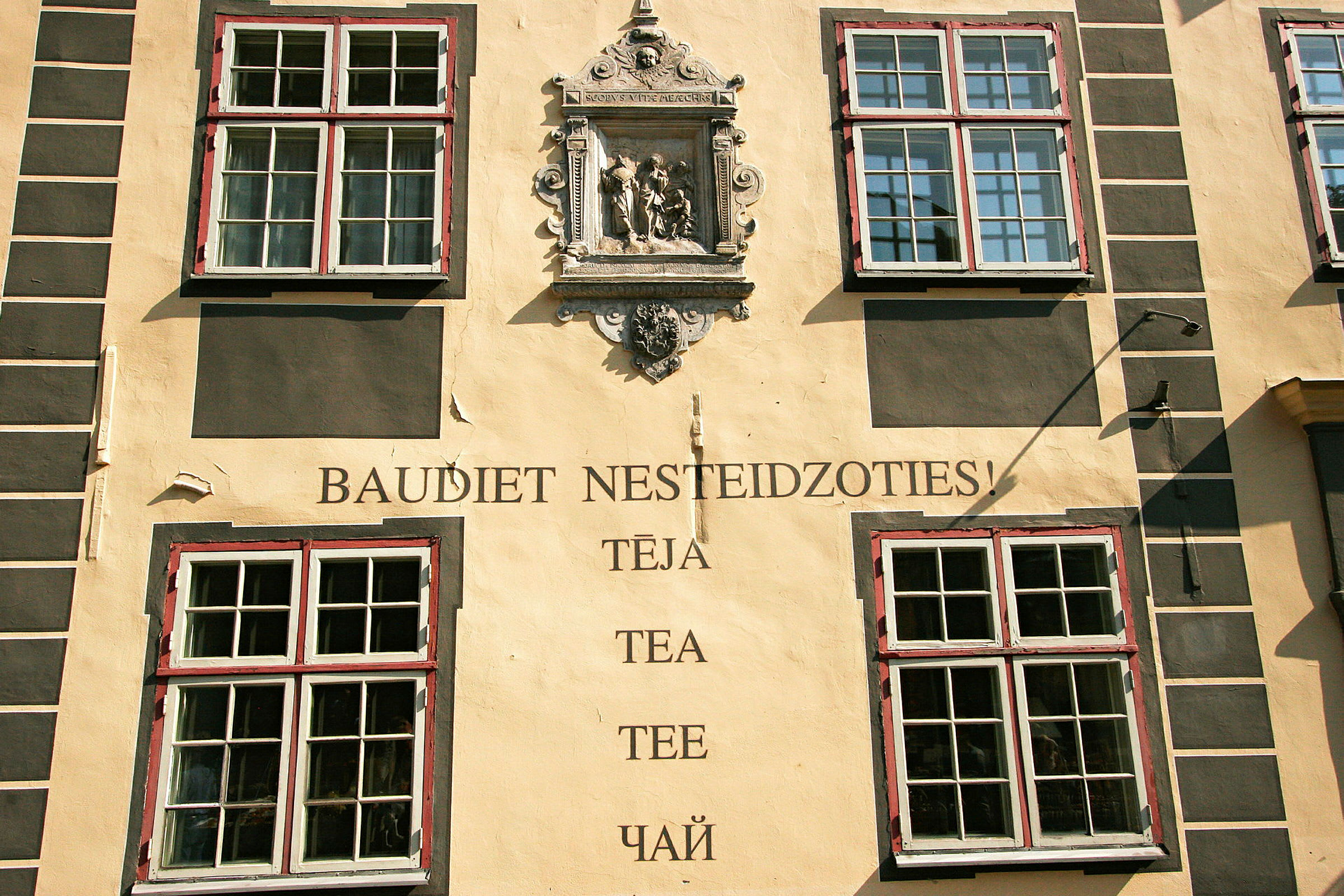 Rakennuksen julkisivu, jossa on neljä suurta ikkunaa, koristeellinen kivi ja teksti "Baudiet nesteidzoties! Tēja, Tea, Tee, Чай". (Tekoälyn luoma teksti)