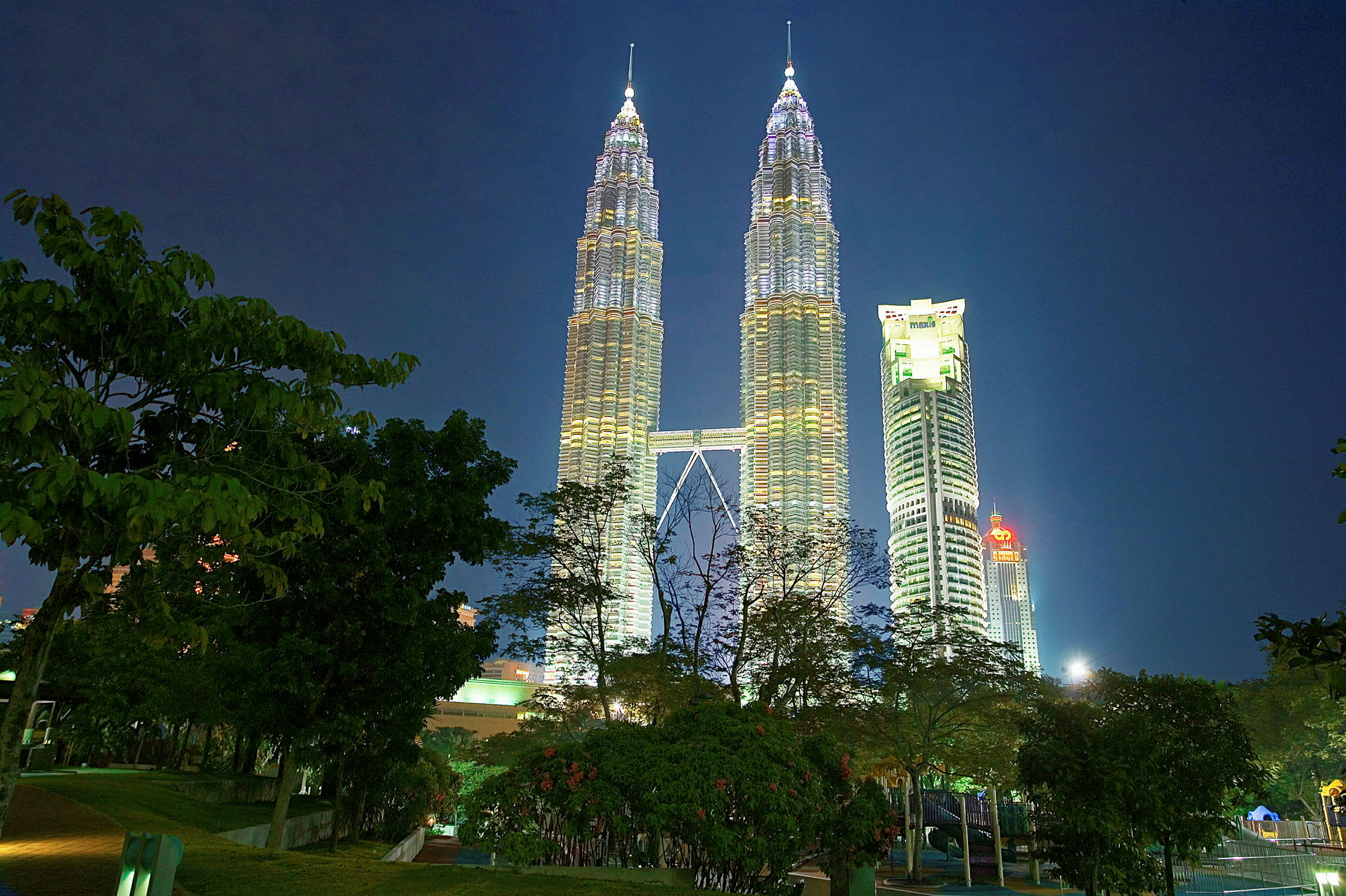 Petronas Twin Towers valaistuna yöllä, edustalla puita. (Tekoälyn luoma teksti)