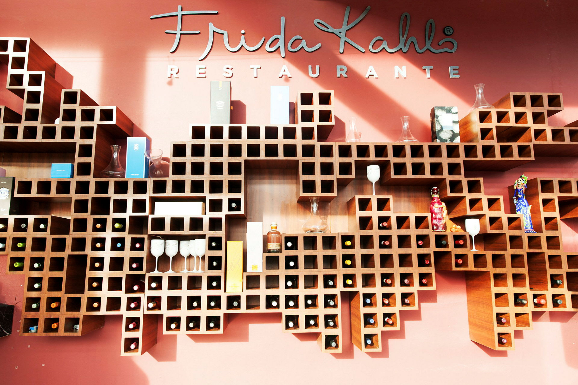 Seinähyllyt ruutukuvioinnilla, täynnä pulloja, laseja ja koriste-esineitä, tekstin "Frida Kahlo Restaurante" edessä. (Tekoälyn luoma teksti)