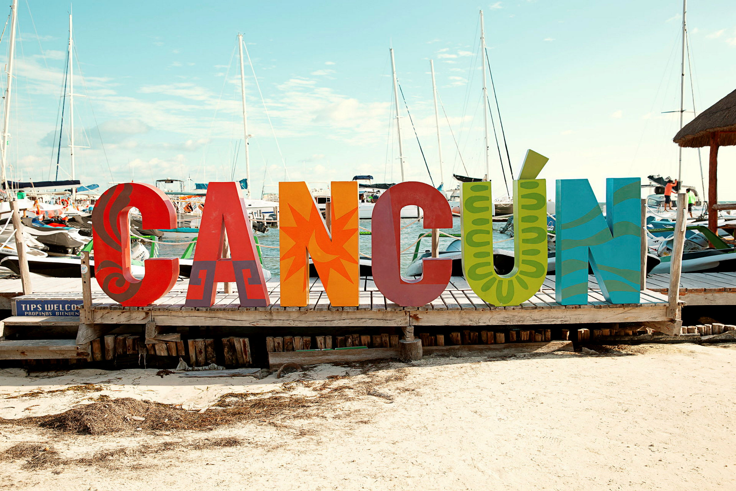 Värikkäitä kirjainveistoksia, jotka muodostavat sanan 'Cancún', seisovat laiturilla veneiden ja mastojen taustalla. (Tekoälyn luoma teksti)