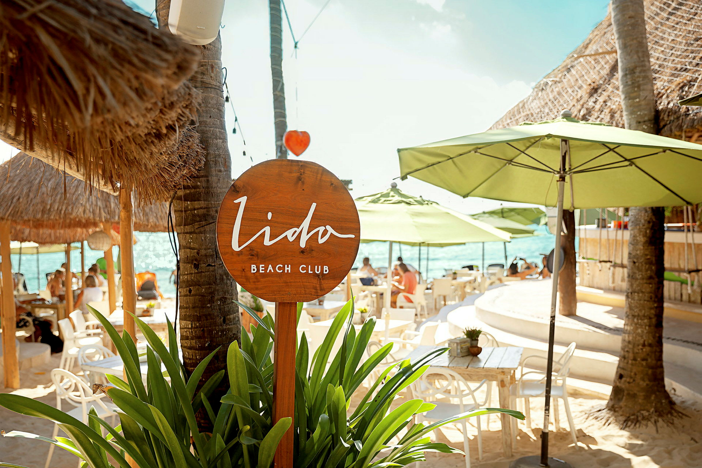 Lido Beach Club -kyltti, jota ympäröivät vihreät kasvit ja aurinkovarjot rannalla. (Tekoälyn luoma teksti)