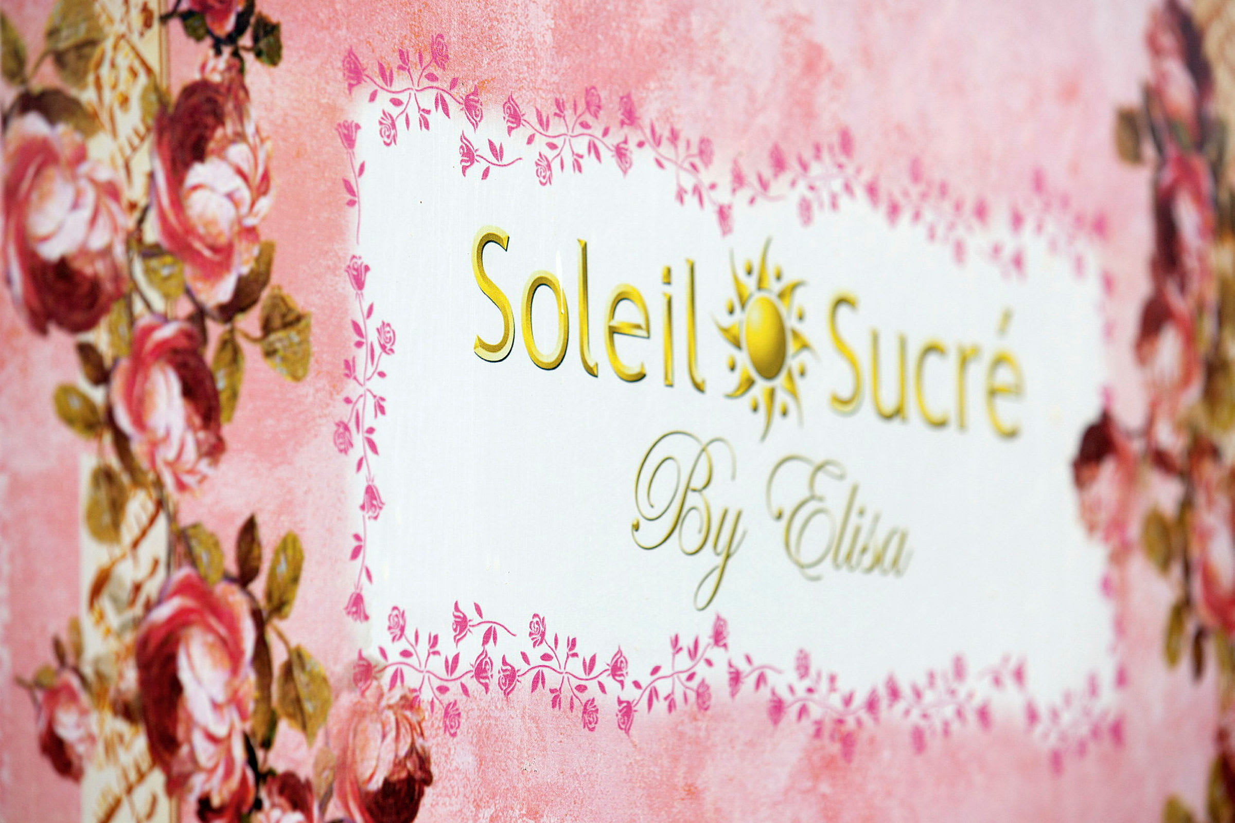 Teksti 'Soleil Sucré By Elisa' koristeellisella taustalla, jossa on vaaleanpunaisia ja kukkaisia yksityiskohtia. (Tekoälyn luoma teksti)