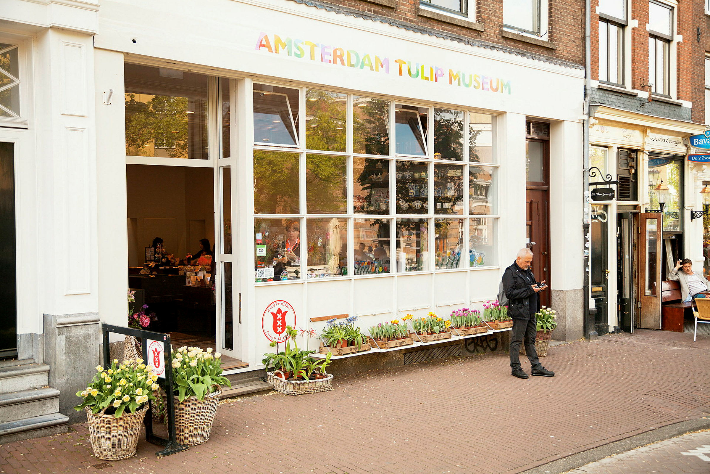 Amsterdamin tulppaanimuseon sisäänkäynti, edessä kukkia ja sisällä ihmisiä. (Tekoälyn luoma teksti)