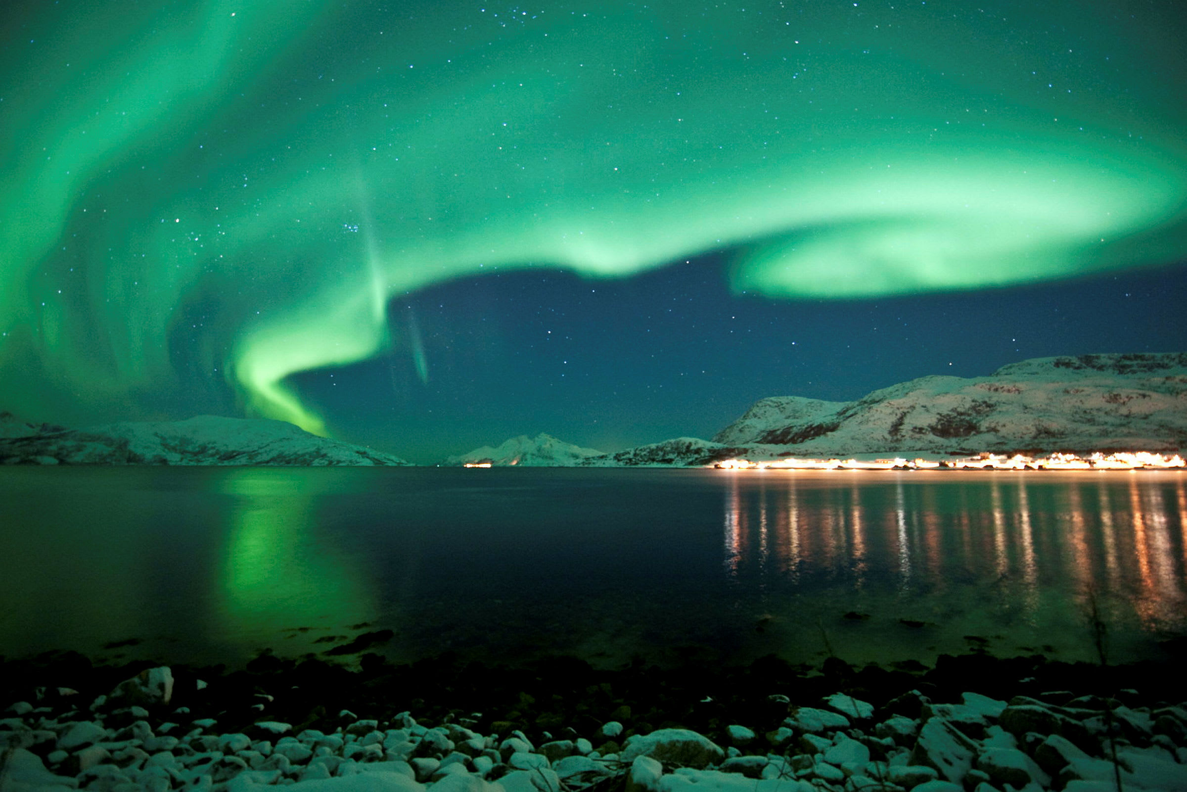 Kuva luonnonilmiöstä, jossa revontulet näkyvät lumipeitteisten vuorten, veden ja valaistun rannikon yllä. (Tekoälyn luoma teksti)