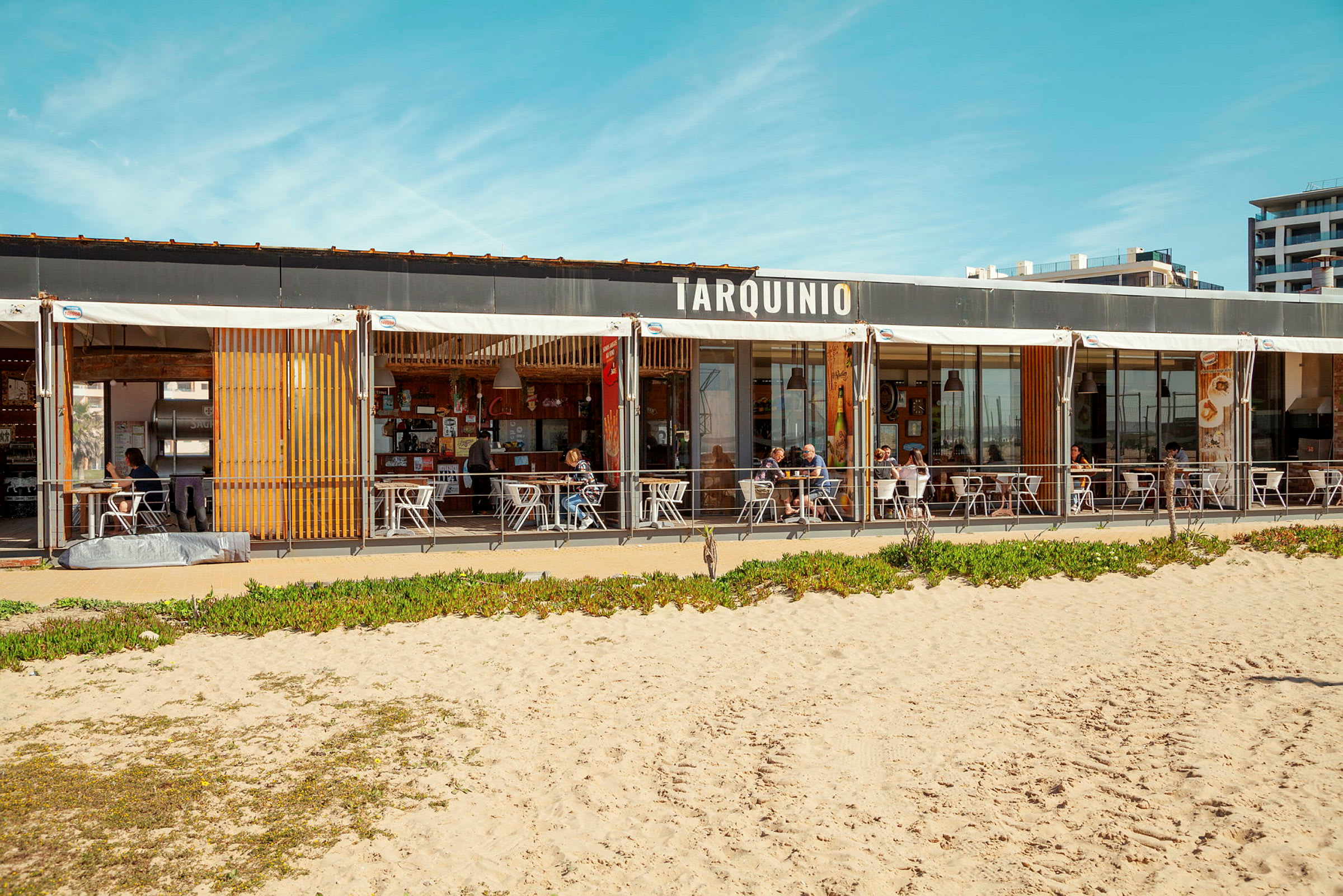 Ravintola rannalla, jossa ulkona istumapaikkoja ja hiekkaranta etualalla, kyltissä lukee 'Tarquinio'. (Tekoälyn luoma teksti)