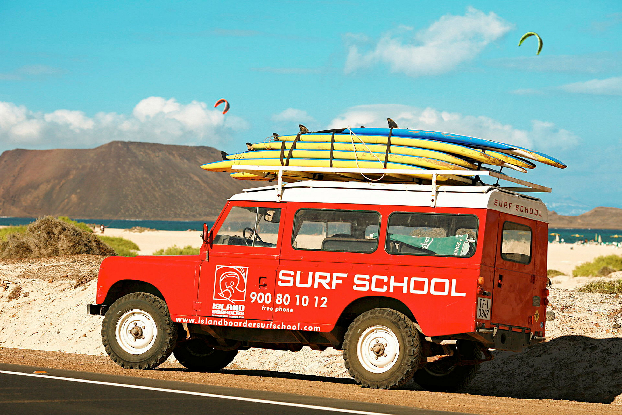 Punainen "Surf School" -tekstitetty auto on pysäköity rannalle, ja sen katolla on surffilautoja. (Tekoälyn luoma teksti)