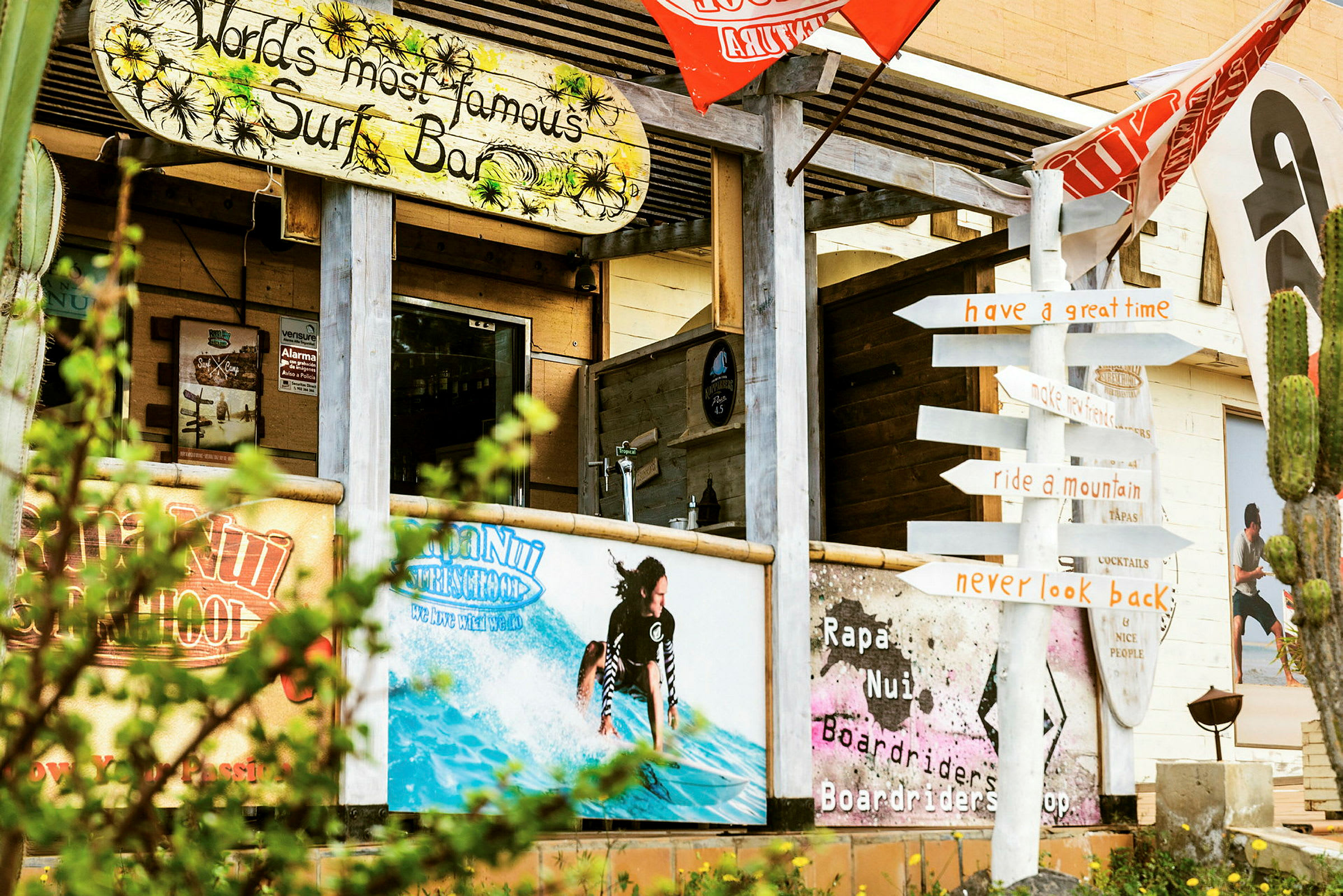 Surffihotelli, jossa on värikkäät kyltit ja koristeet, mukaan lukien hahmo surffaamassa vedessä, sekä tienviitta, joka osoittaa eri suuntiin. (Tekoälyn luoma teksti)