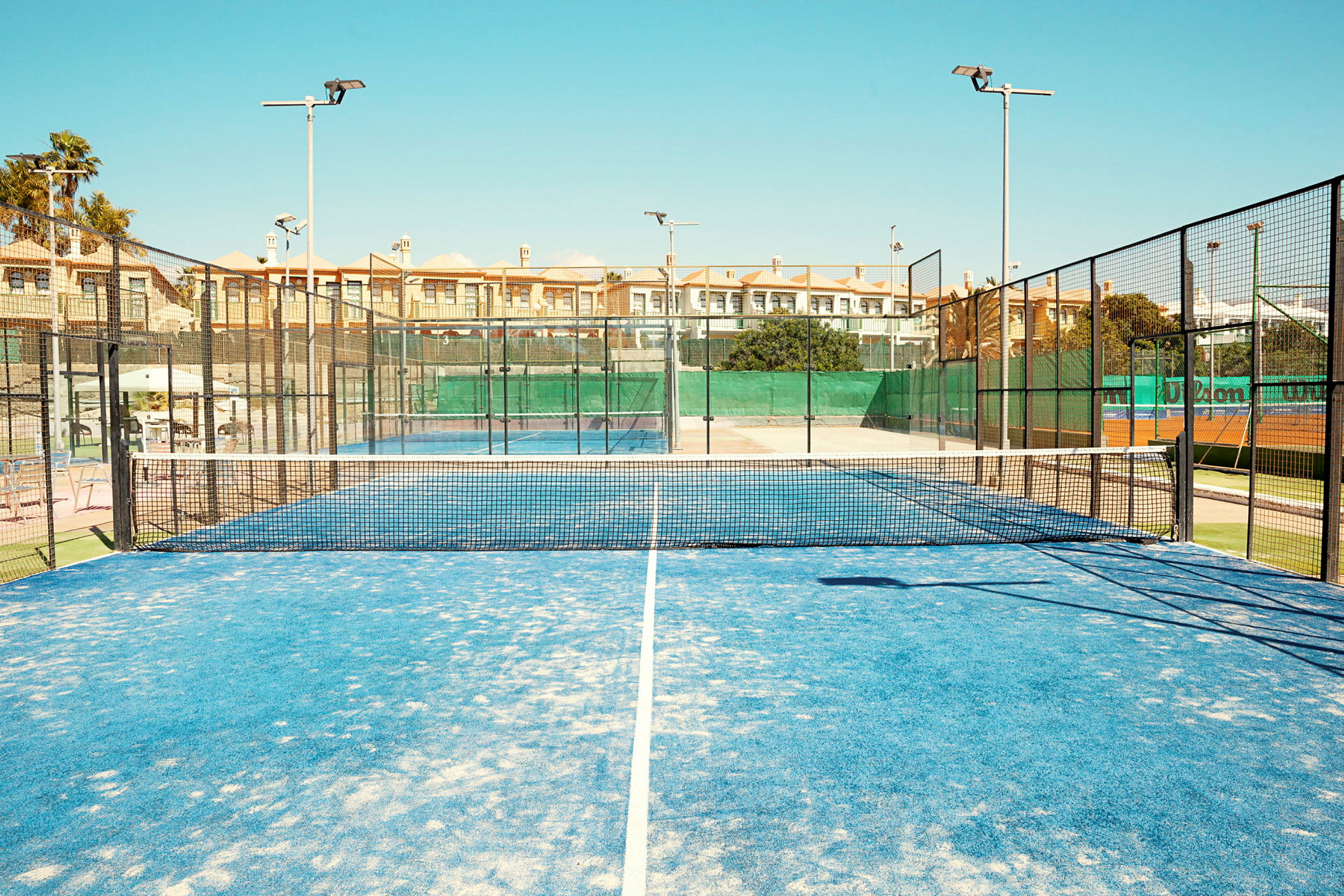 Padel-kenttä, jossa sininen alusta ja verkko keskellä, ympärillä aidat ja taustalla rakennuksia. (Tekoälyn luoma teksti)