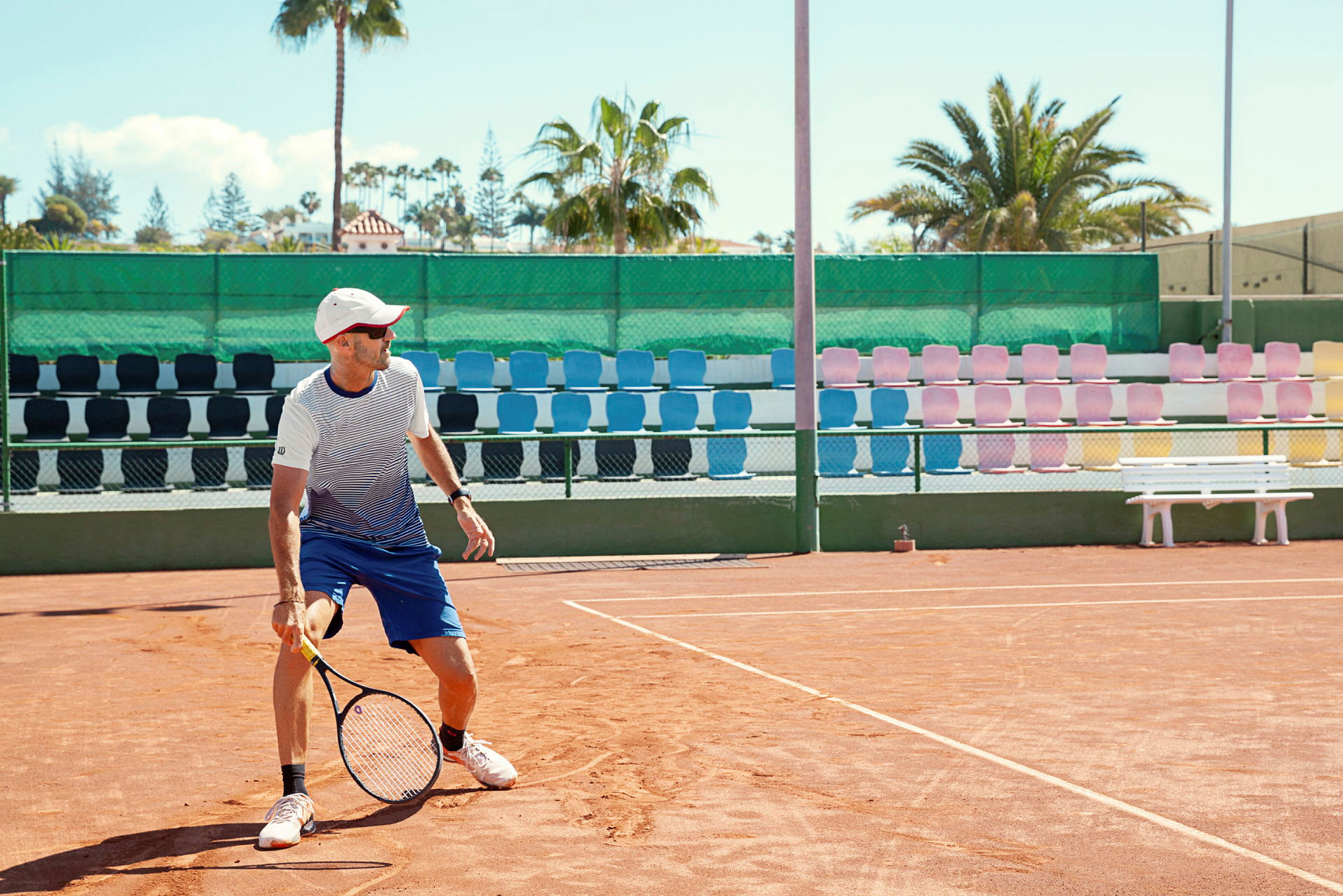 Tenniskenttä, jossa pelaaja pitää mailaa. Taustalla värikkäitä istuimia ja palmuja. (Tekoälyn luoma teksti)