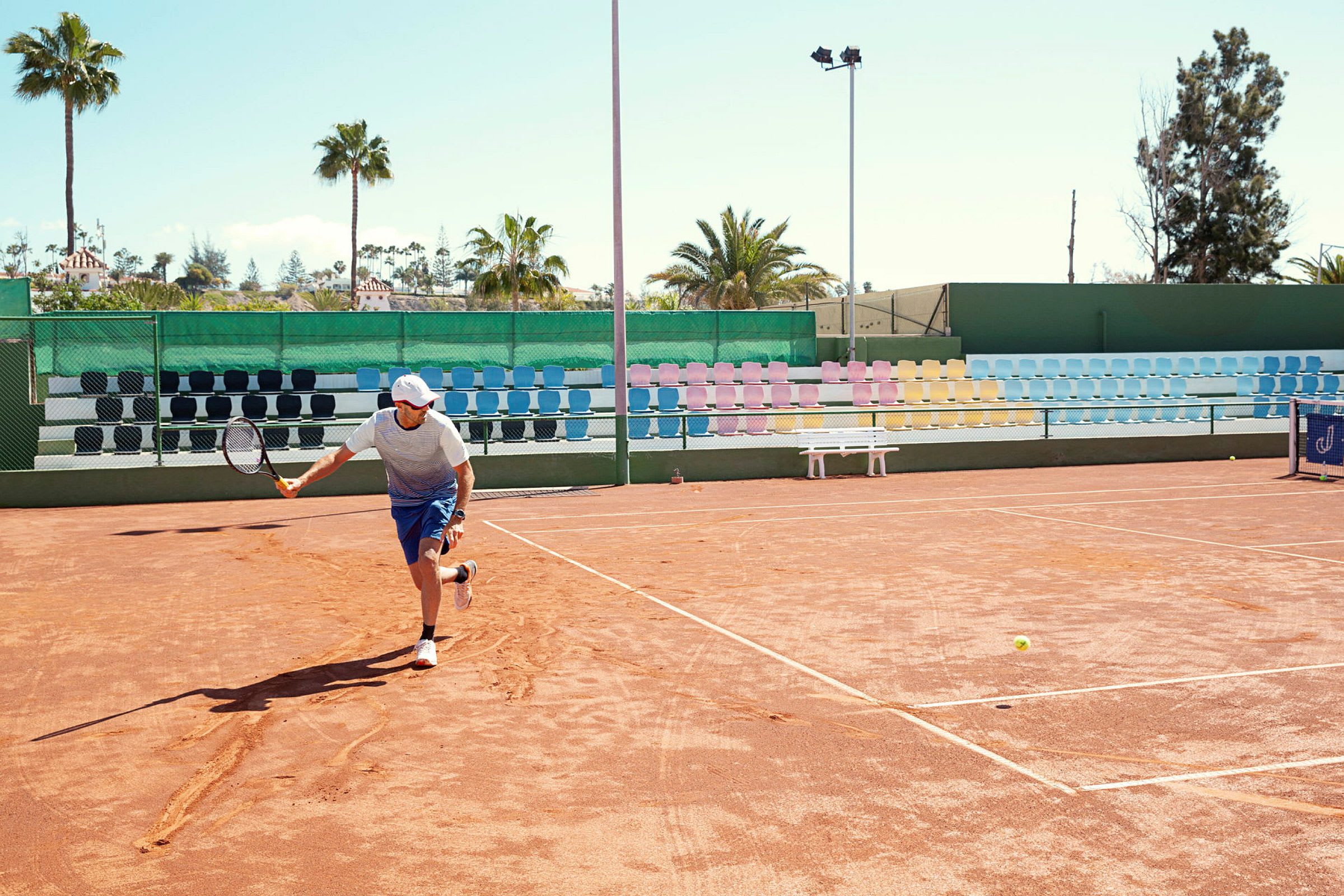 Henkilö pelaa tennistä ulkona hiekkakentällä, taustalla värikkäitä katsomotuoleja ja palmuja. (Tekoälyn luoma teksti)