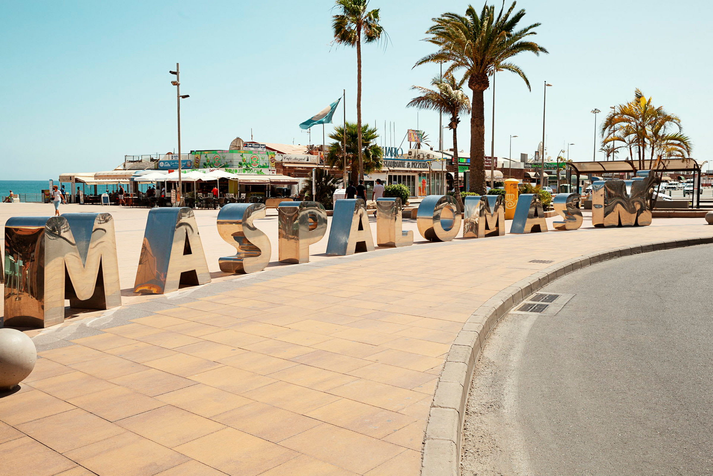 Suuret, kiiltävät kirjaimet muodostavat sanan "MASPALOMAS" jalkakäytävällä aukion läheisyydessä, jossa on palmuja. (Tekoälyn luoma teksti)