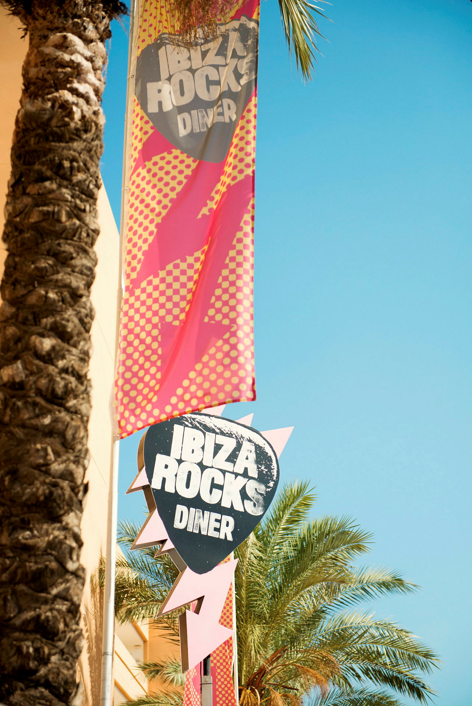 Ibiza Rocks Dinerin kyltti ja banneri, ympärillä palmuja ja selkeä taivas. (Tekoälyn luoma teksti)