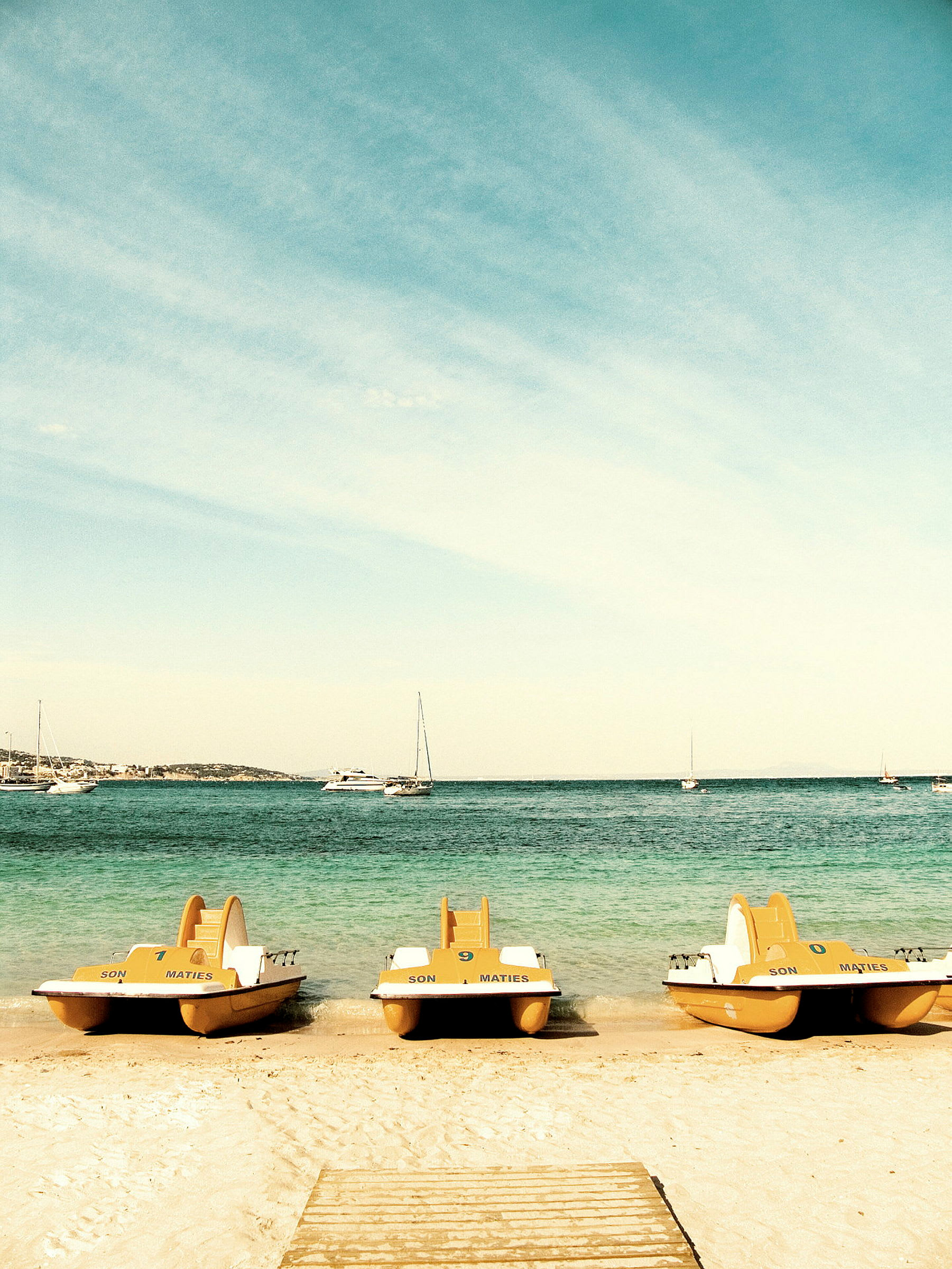Kolme polkuvenettä on pysäköity hiekkarannalle, taustalla meri ja purjeveneitä. (Tekoälyn luoma teksti)