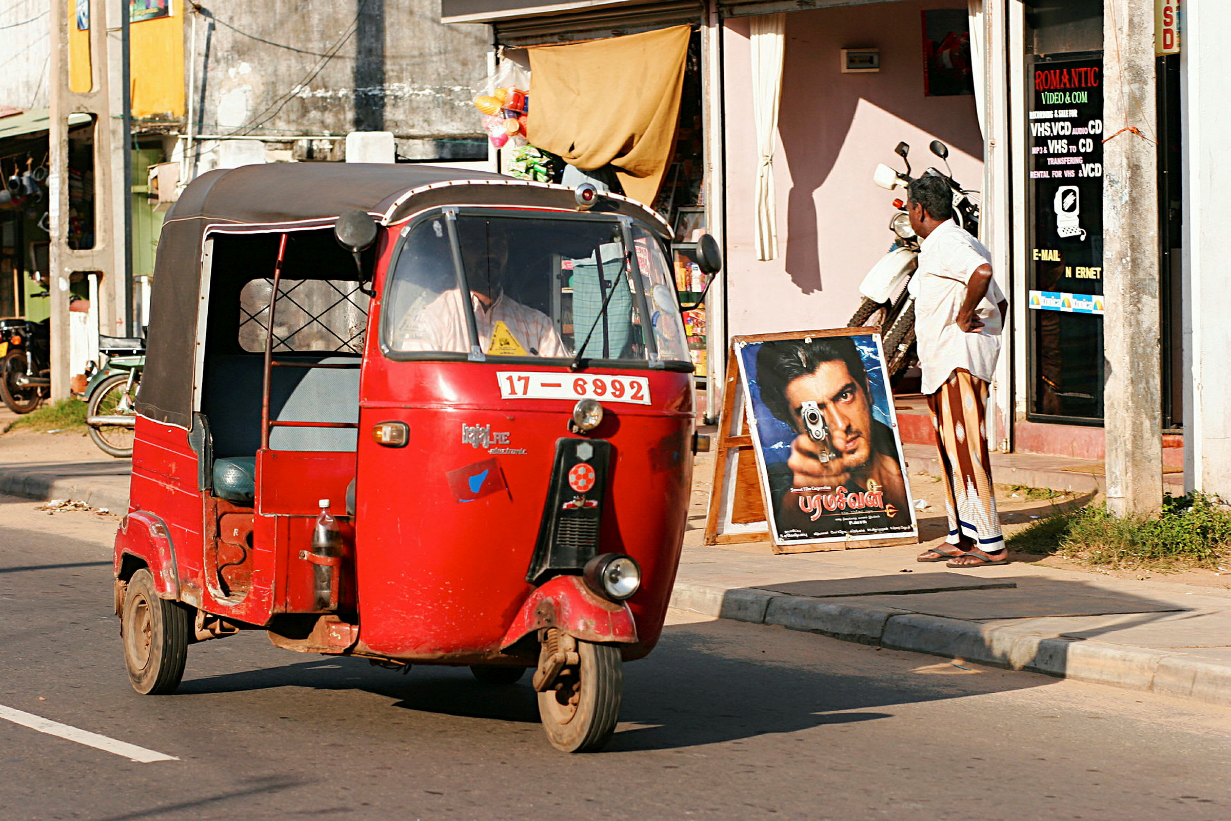 Punainen tuk-tuk ajaa ohi kaupan aurinkoisella kadulla. (Tekoälyn luoma teksti)