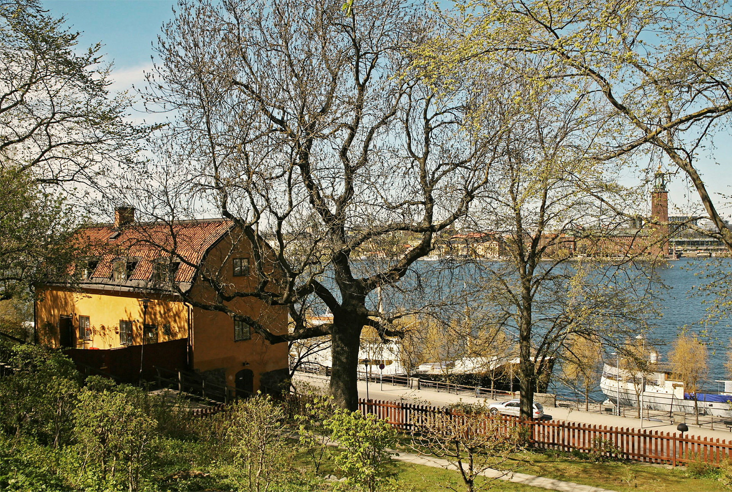 Keltainen rakennus, jossa on punainen katto, sijaitsee tien vieressä, ja etualalla on puita. Taustalla näkyy järvi. (Tekoälyn luoma teksti)