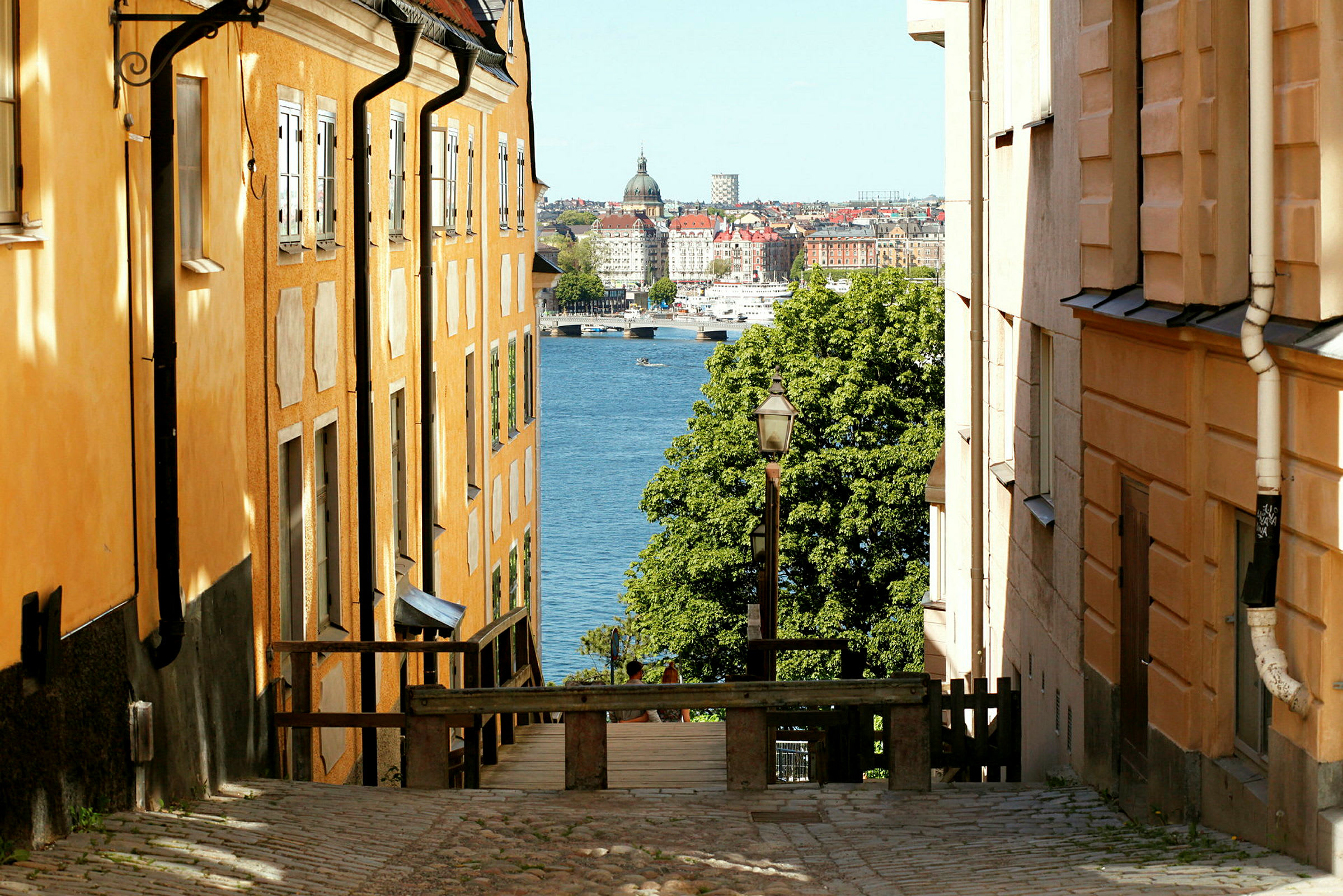 Kuva kadusta, jossa on oranssinvärisiä rakennuksia kummallakin puolella, viettäen alas kohti vettä ja taustan kaupunkia. (Tekoälyn luoma teksti)