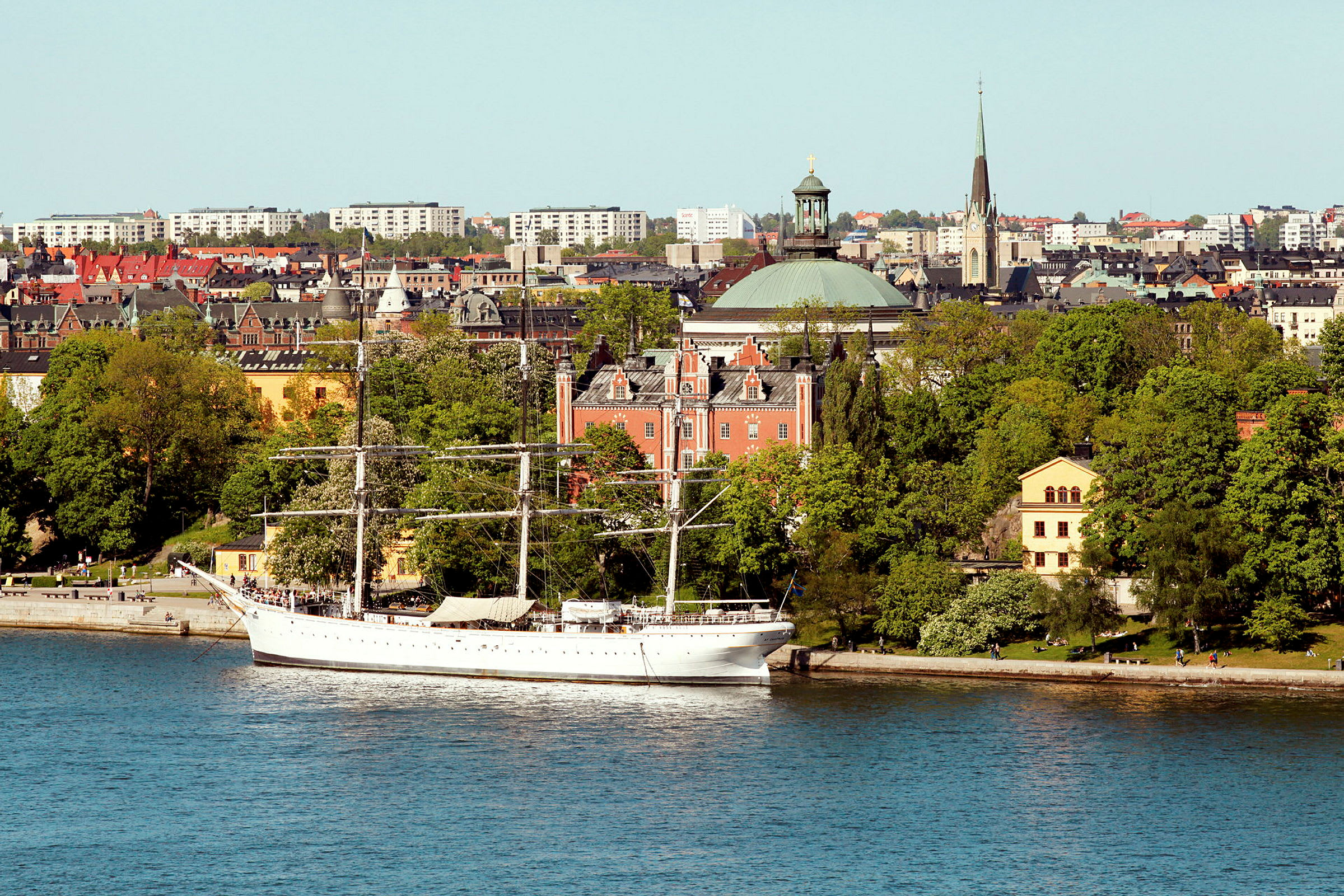 Valkoinen purjelaiva on ankkuroituna kaupungin rantaan veden äärellä. Taustalla on puita, useita vanhoja rakennuksia ja kaupungin rakenteita. (Tekoälyn luoma teksti)