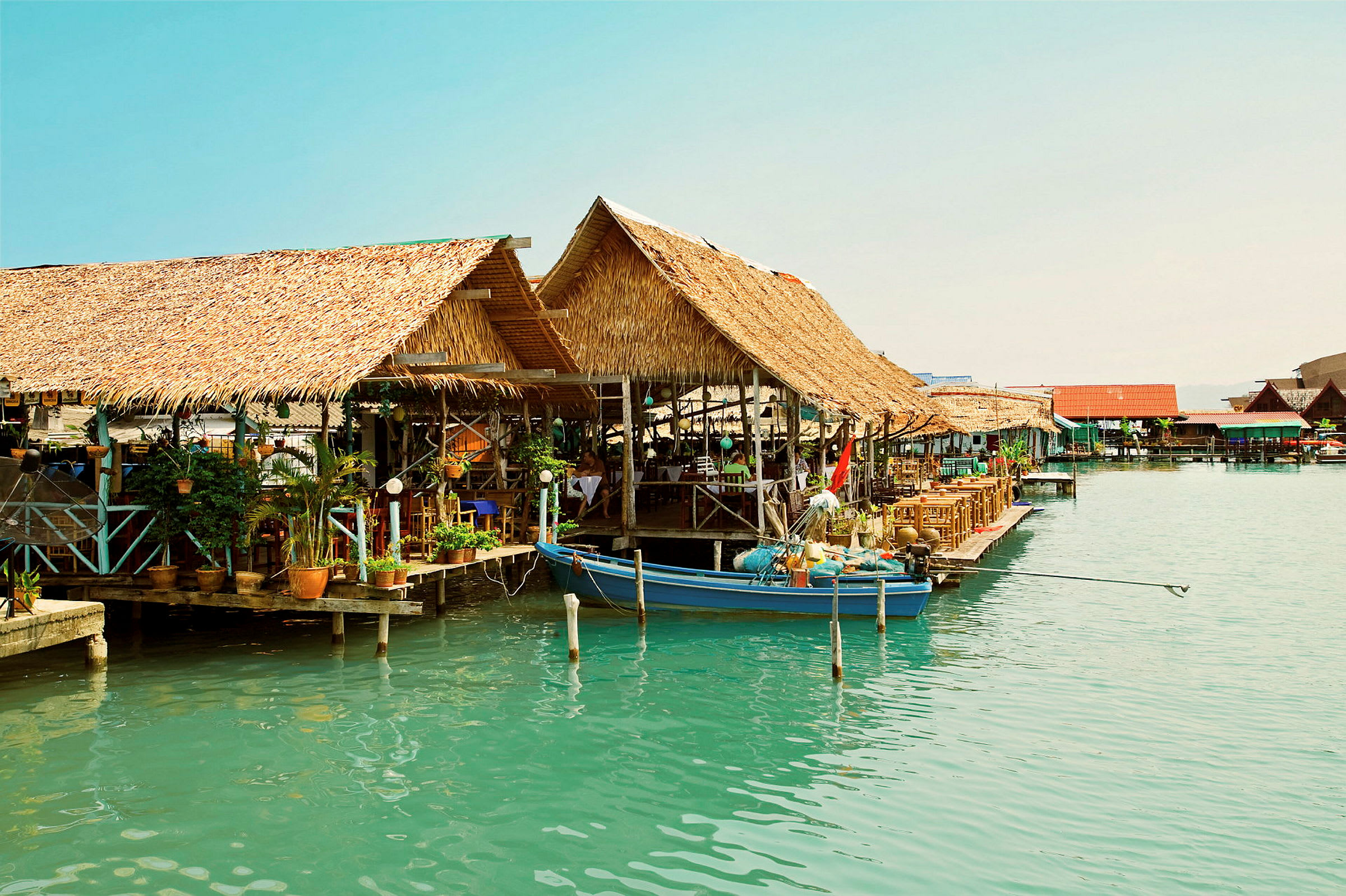 Useita vesirannan olkikattoisia rakennuksia, joista yksi on veneen vieressä. (Tekoälyn luoma teksti)