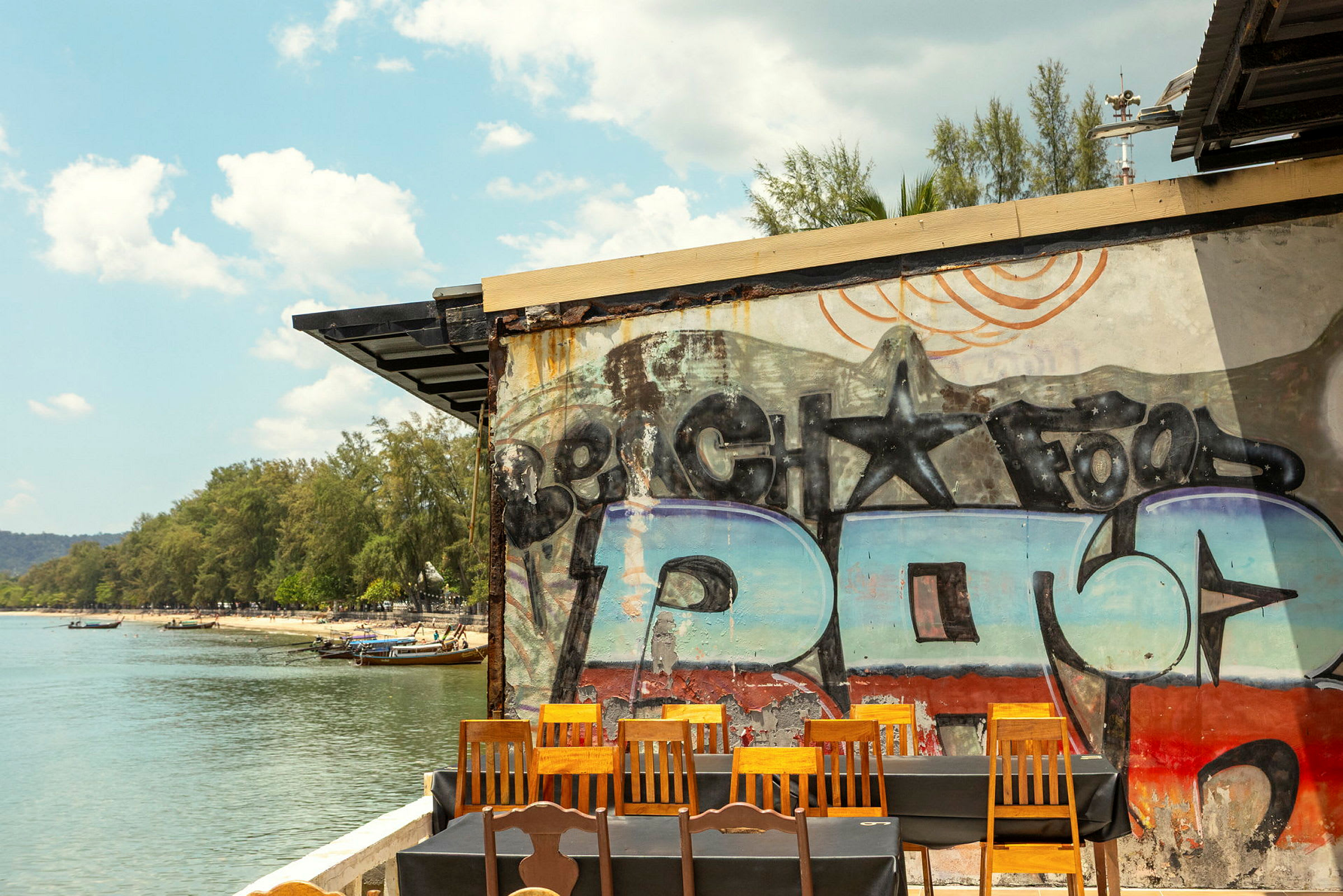 Näkymä rannalle, jossa on vettä, puita ja veneitä. Kuvan etualalla näkyy seinä graffitilla sekä tuoleja ja pöytiä. (Tekoälyn luoma teksti)