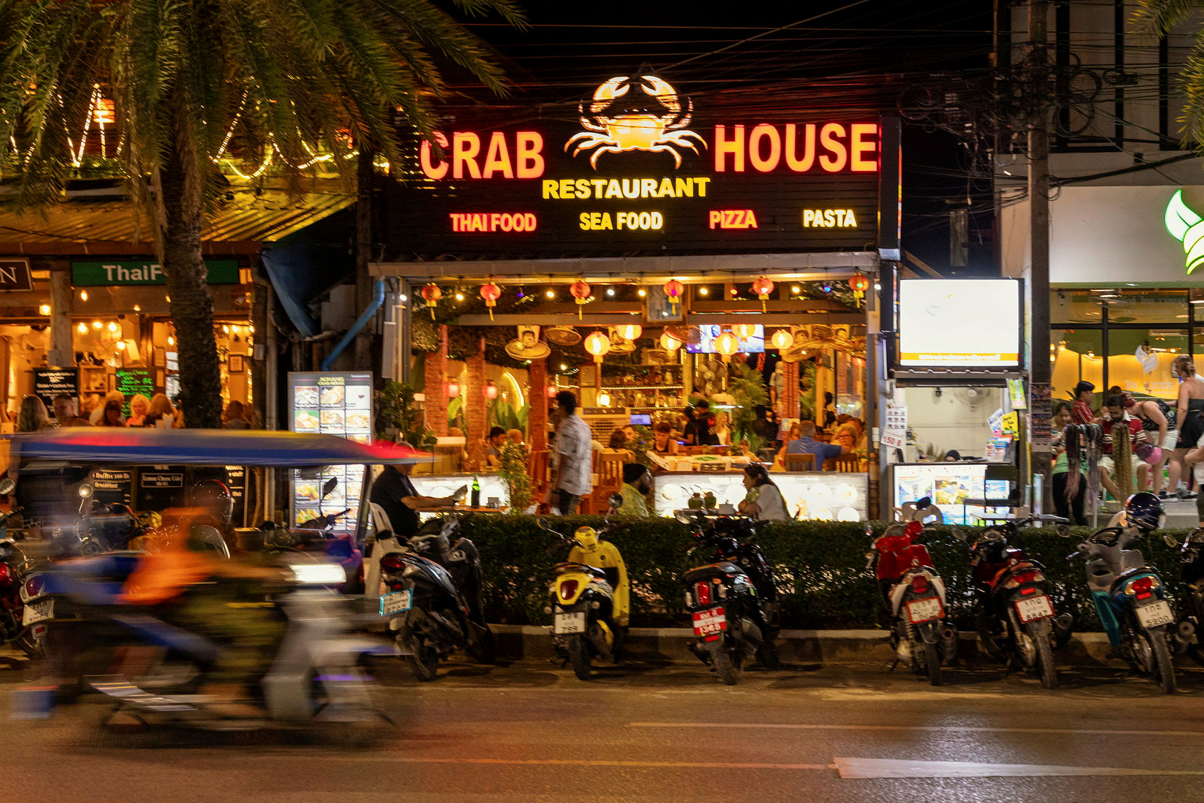 Kuva vilkkaalta kadulta, jossa Crab House -ravintola on keskipisteenä. Useita moottoripyöriä on pysäköity ravintolan eteen, ja moottoripyörä ohittaa pyöräilijän kanssa. (Tekoälyn luoma teksti)