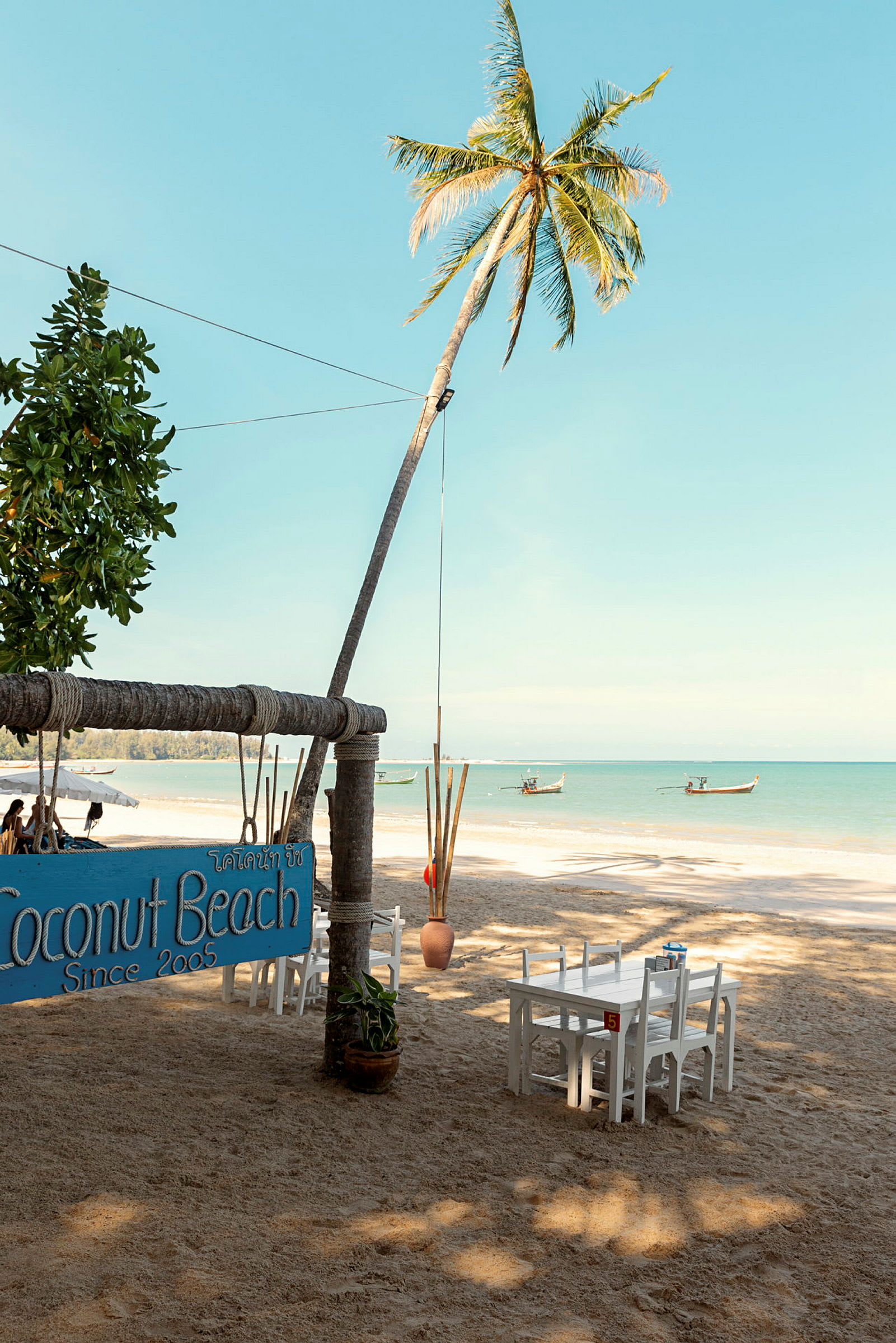 Ranta palmuineen, pöytineen ja tuoleineen, sekä kyltti, joka osoittaa Coconut Beach. (Tekoälyn luoma teksti)