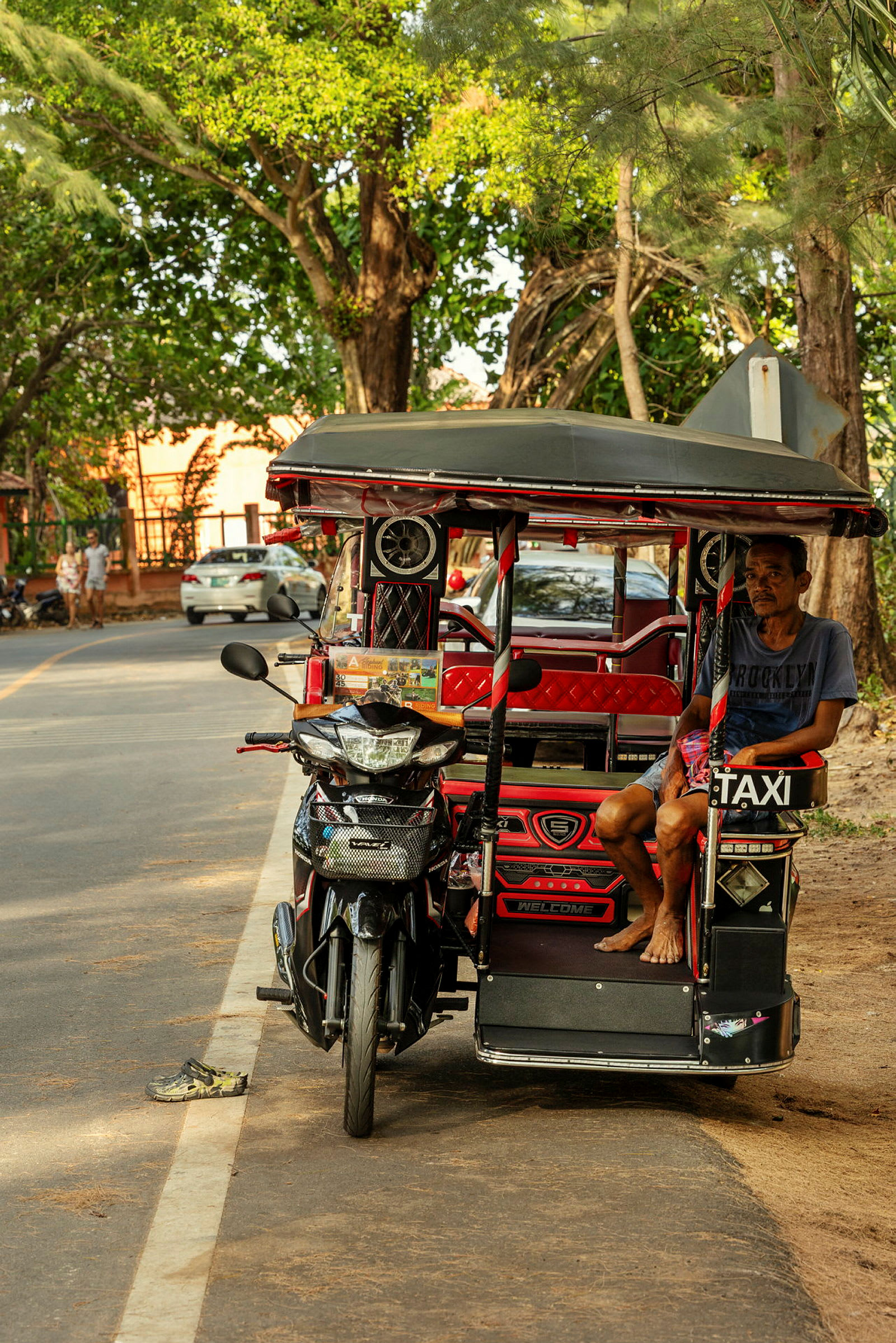 Tuk-tuk on pysäköity kadun varrelle. Mies istuu ajoneuvossa, ympärillä on puita ja muutamia pysäköityjä autoja. (Tekoälyn luoma teksti)