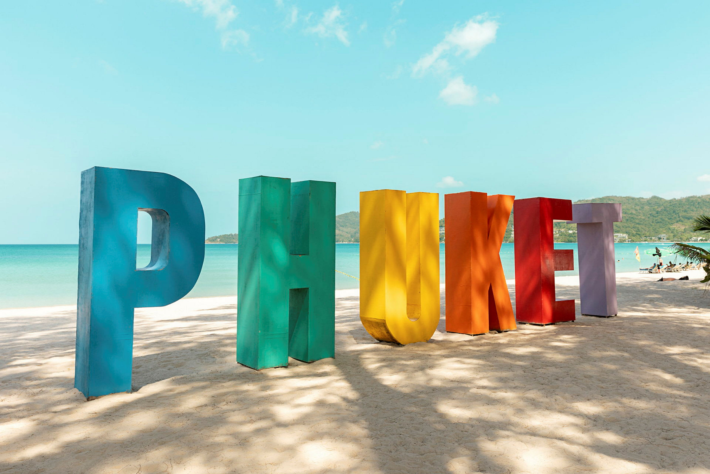 Värikkäät kirjaimet muodostavat sanan 'Phuket' hiekkarannalla, taustalla näkyvät meri ja taivas. (Tekoälyn luoma teksti)