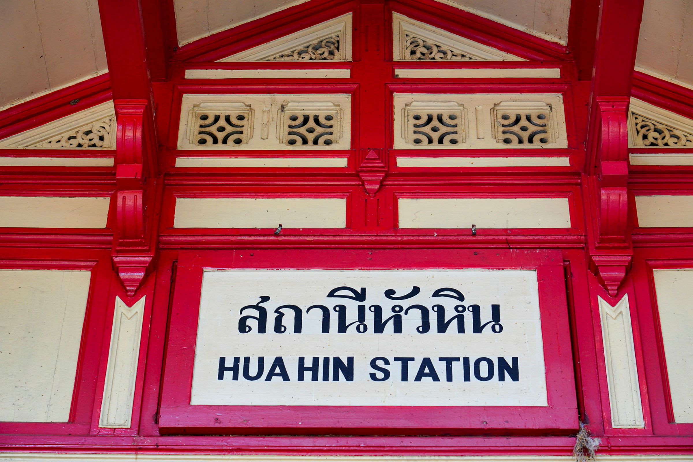Kyltti, jossa lukee 'Hua Hin station', thaimaalaisilla kirjaimilla ja koristeellinen punainen ja valkoinen kehys. (Tekoälyn luoma teksti)